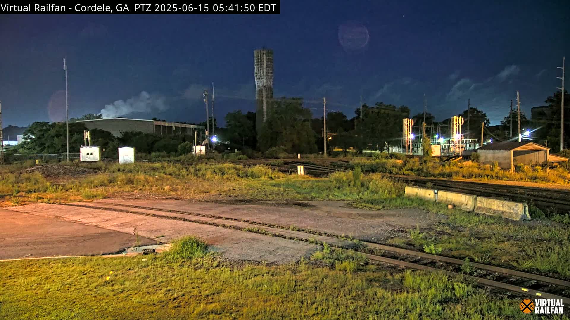 Under a dark, clear night sky, a nighttime view shows railroad tracks near industrial buildings, some with faint smoke rising, and a tall, slender tower.