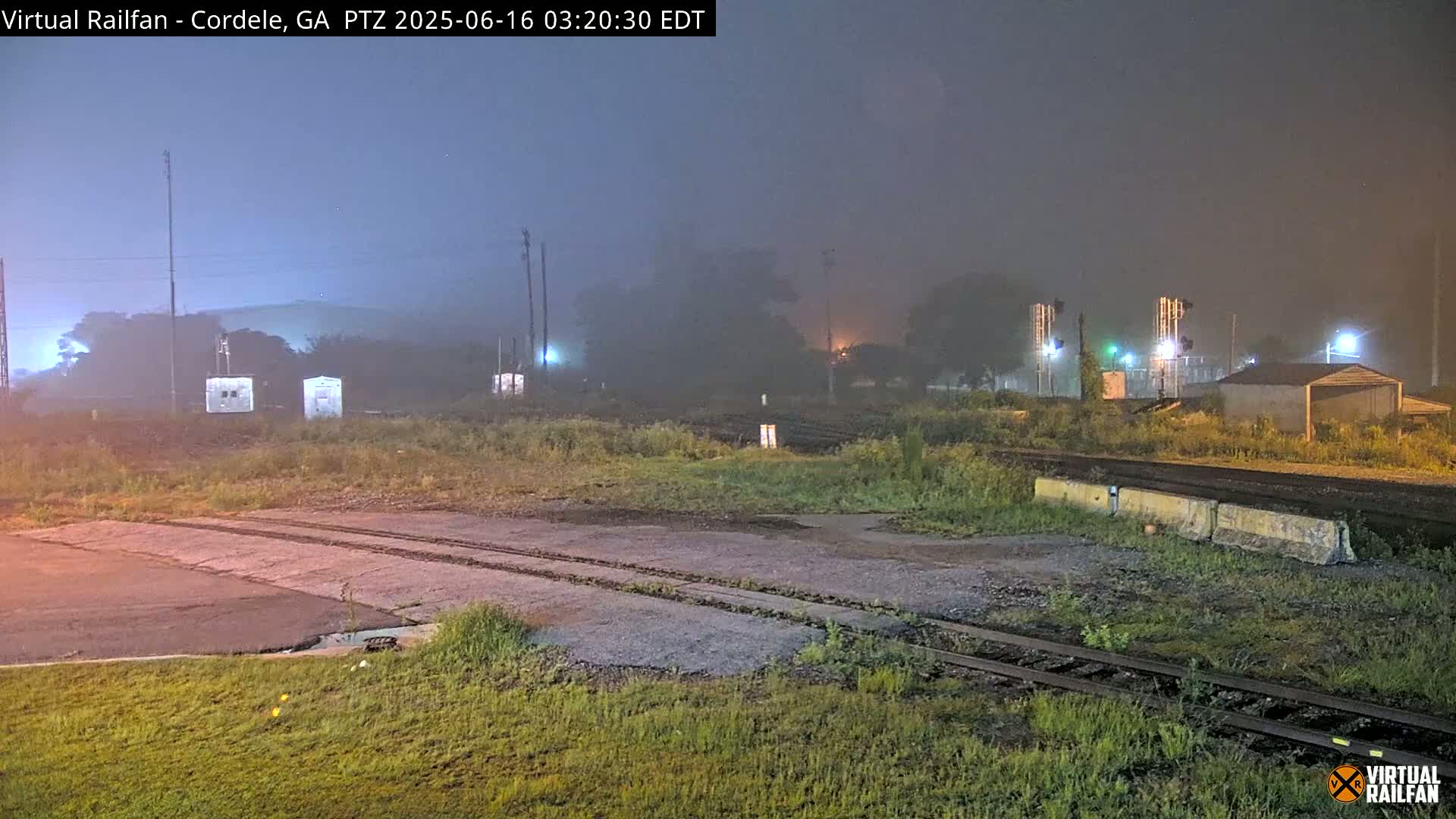 A foggy nighttime view of a railroad yard with several small structures, train tracks, and overgrown vegetation.