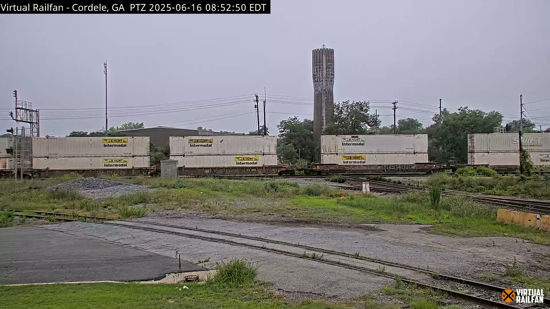 Several stacks of white intermodal shipping containers sit on railroad tracks next to a tall water tower under an overcast sky.
