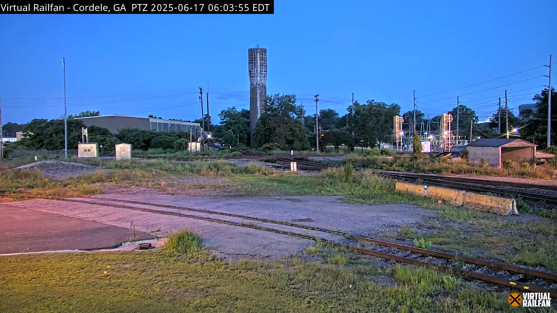 Under a twilight sky, a rail yard with several tracks, a tall water tower, and some industrial buildings is visible.