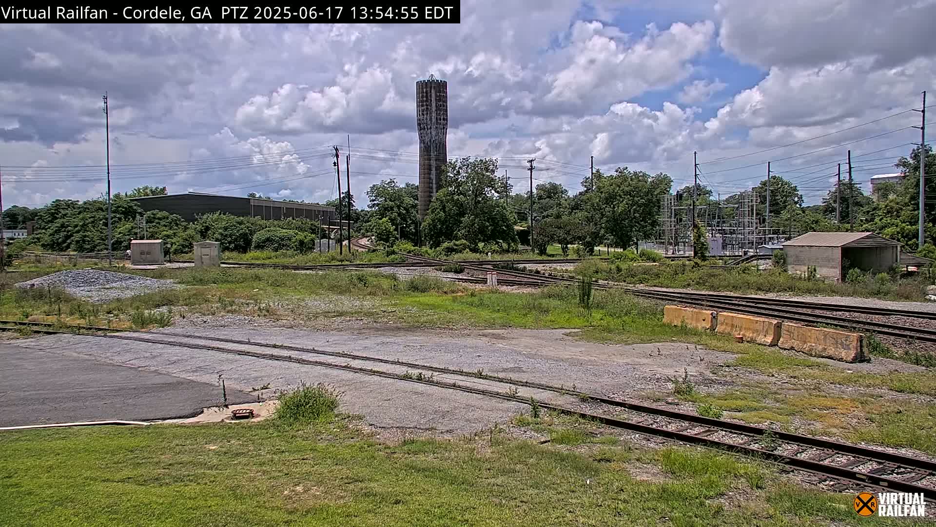 A partly cloudy day reveals a railway switching yard with a tall water tower in the background.