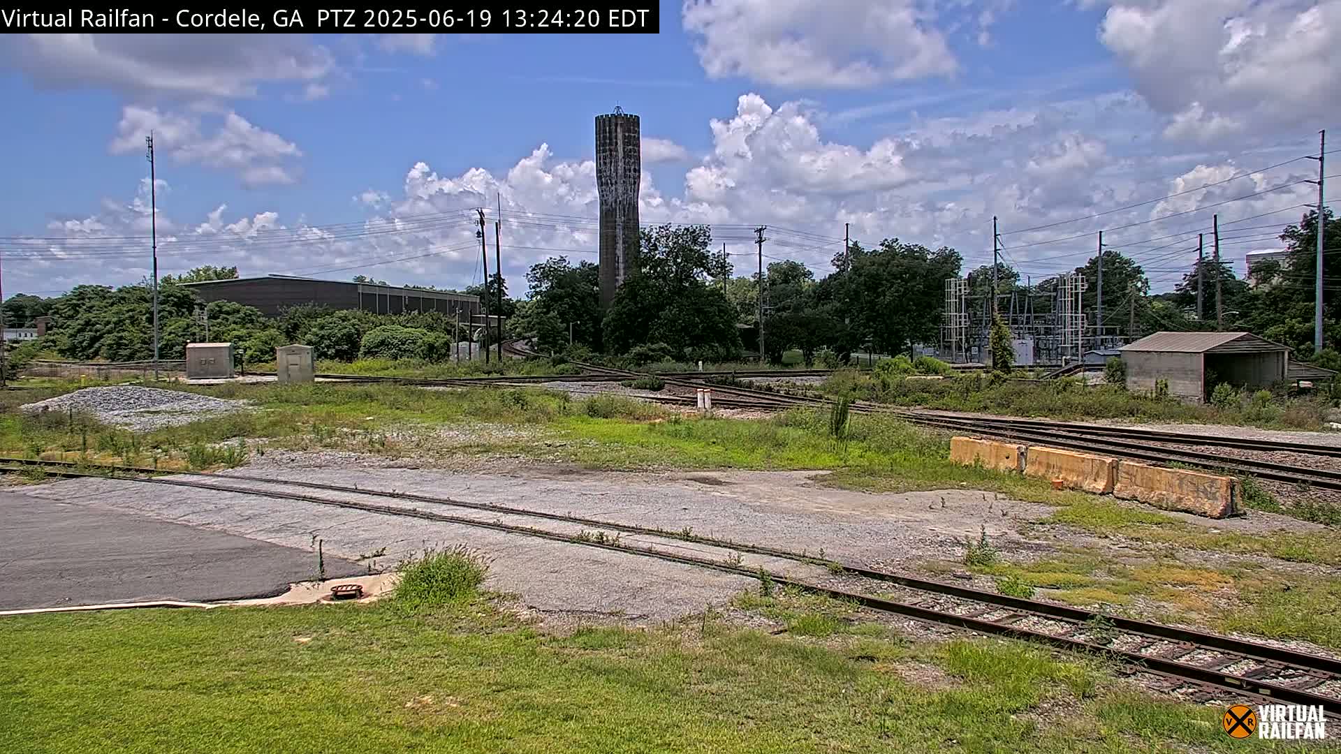 A partly cloudy day reveals a railway switching yard with a tall water tower in the background.