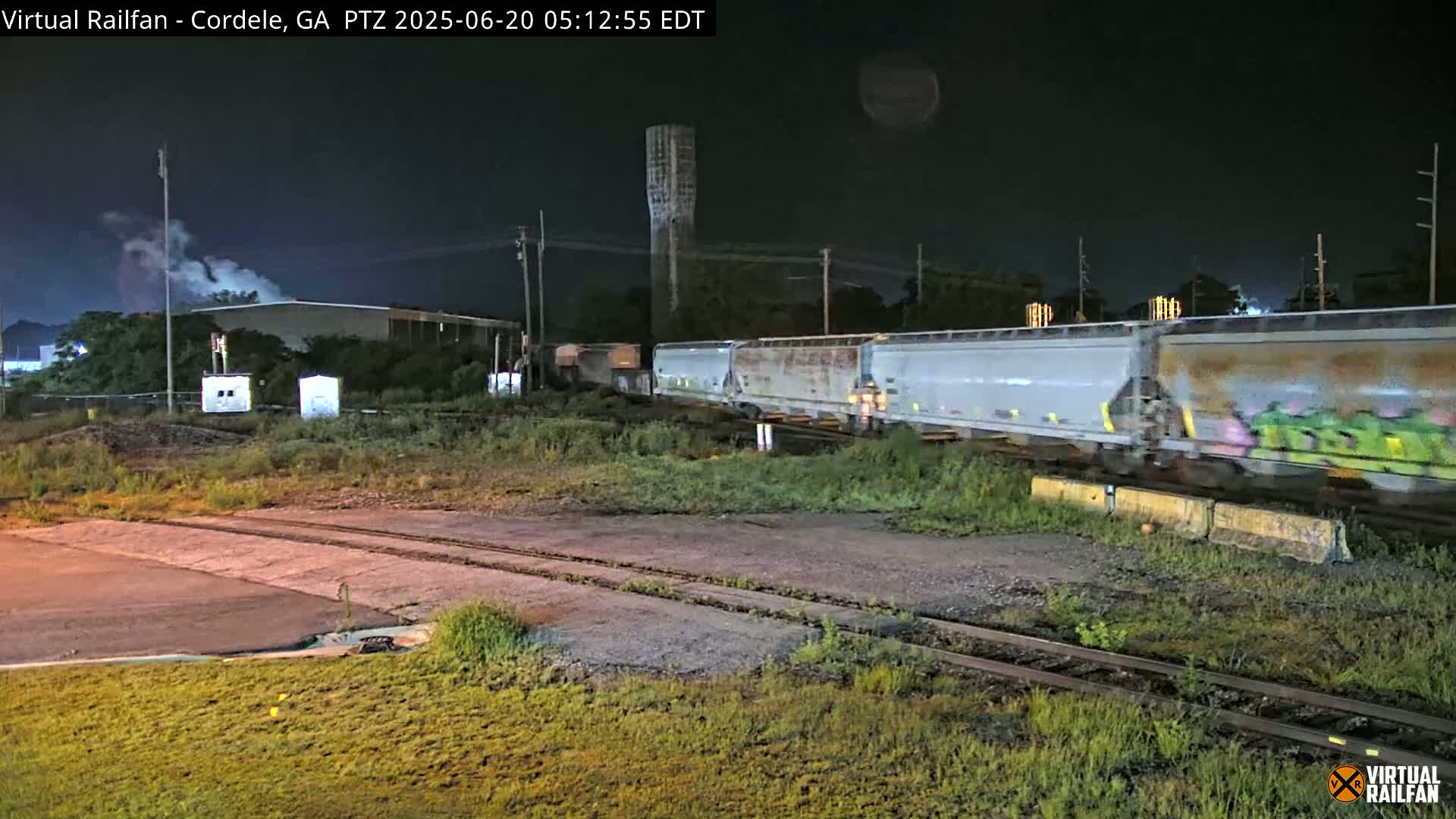 At night, a long freight train passes by a water tower and industrial buildings under a dark sky.