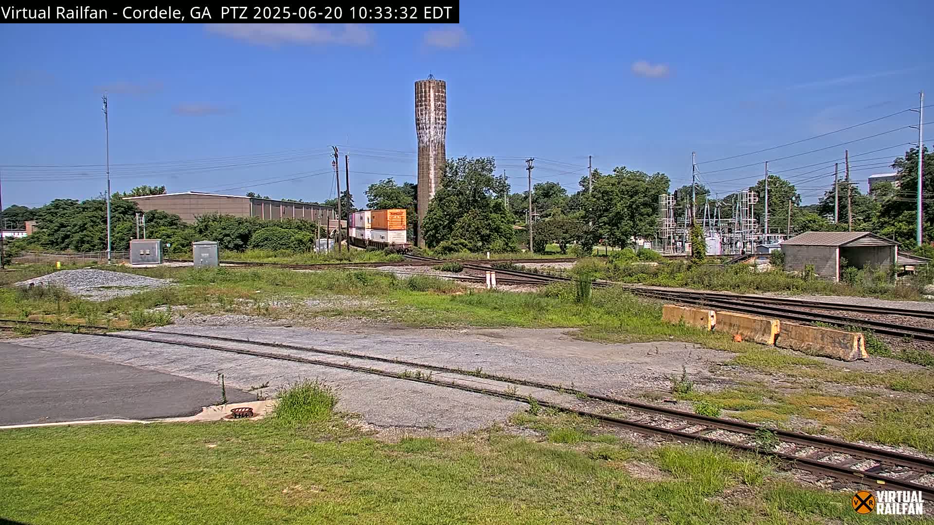 A train car sits near a tall water tower and railroad tracks on a sunny day, with various industrial buildings and vegetation in the background.