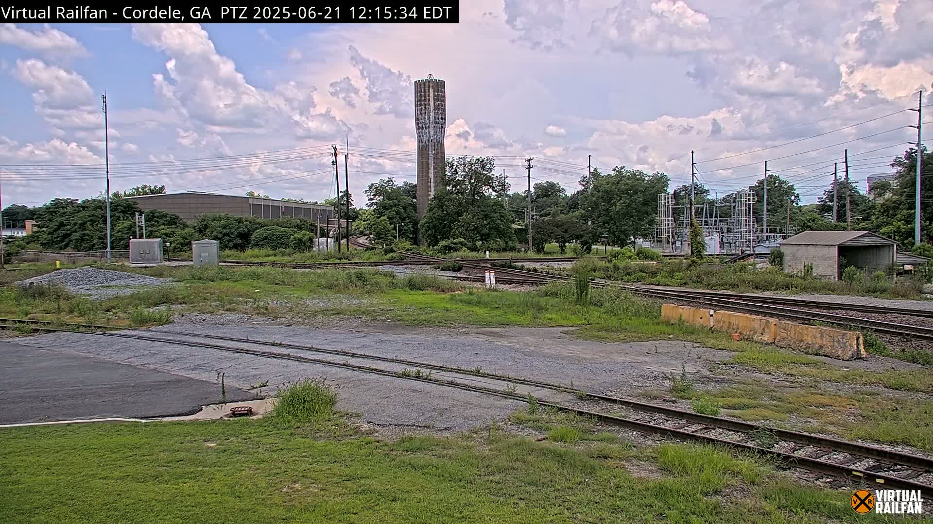 A mostly cloudy sky hangs over a railroad yard with several intersecting tracks, a water tower, and industrial buildings.