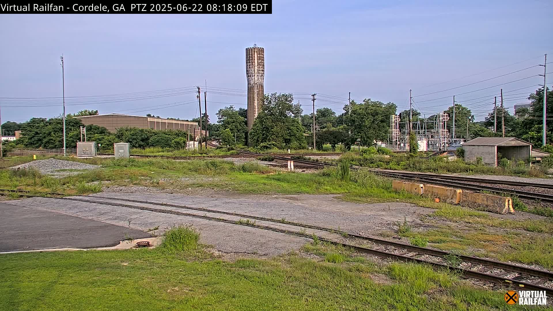 A tall, slender water tower stands in the background of a sunny outdoor scene featuring a railway switching yard with multiple tracks, overgrown vegetation, and industrial buildings.