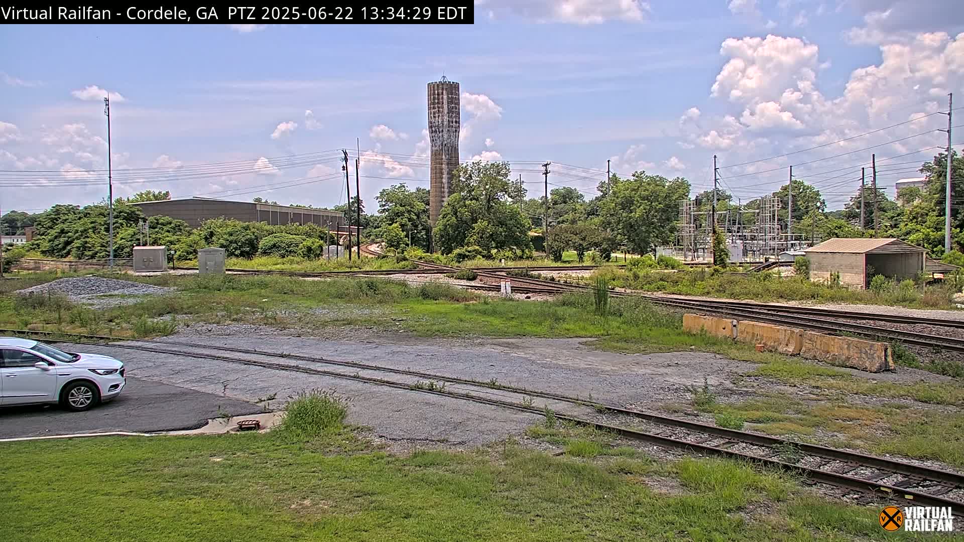 A mostly sunny day shows a view of railroad tracks and switching yards, a tall water tower, and a white SUV parked near a grassy area.
