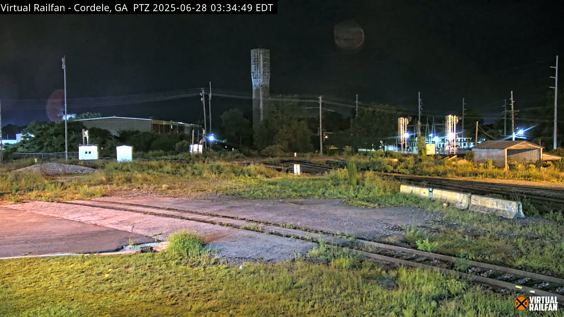 A nighttime view of railroad tracks and industrial buildings, with a water tower visible in the background under a dark sky.