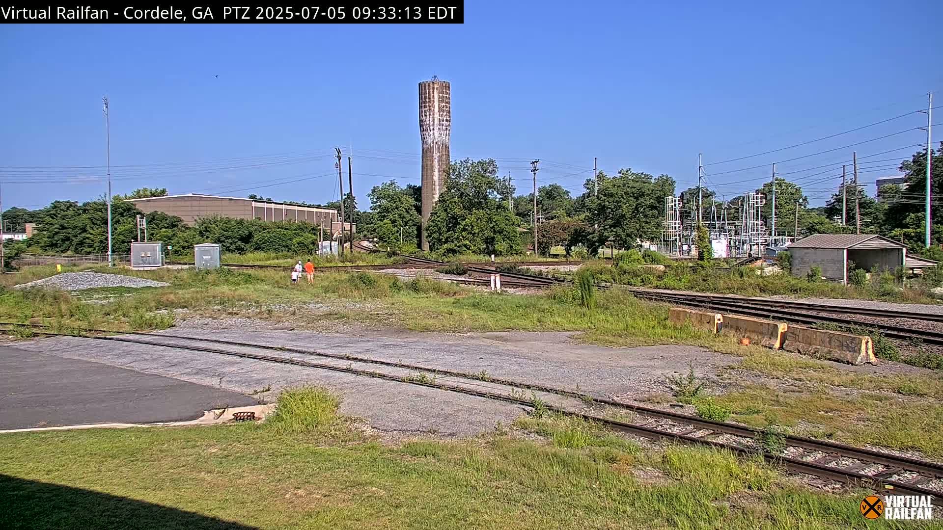 A view of railroad tracks and switching areas on a sunny day, with a water tower and industrial buildings in the background.