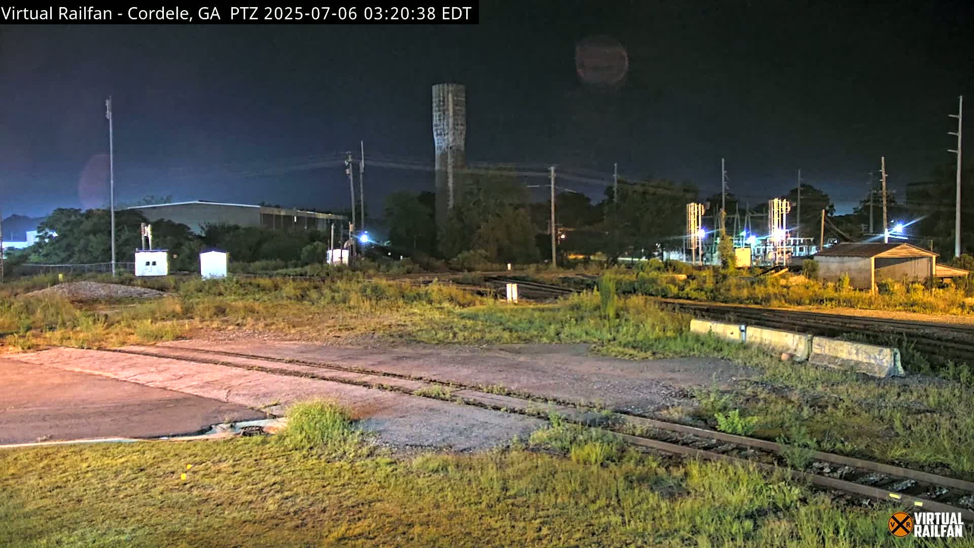 A nighttime view of a railway crossing near industrial buildings and a tall water tower, under a dark sky.