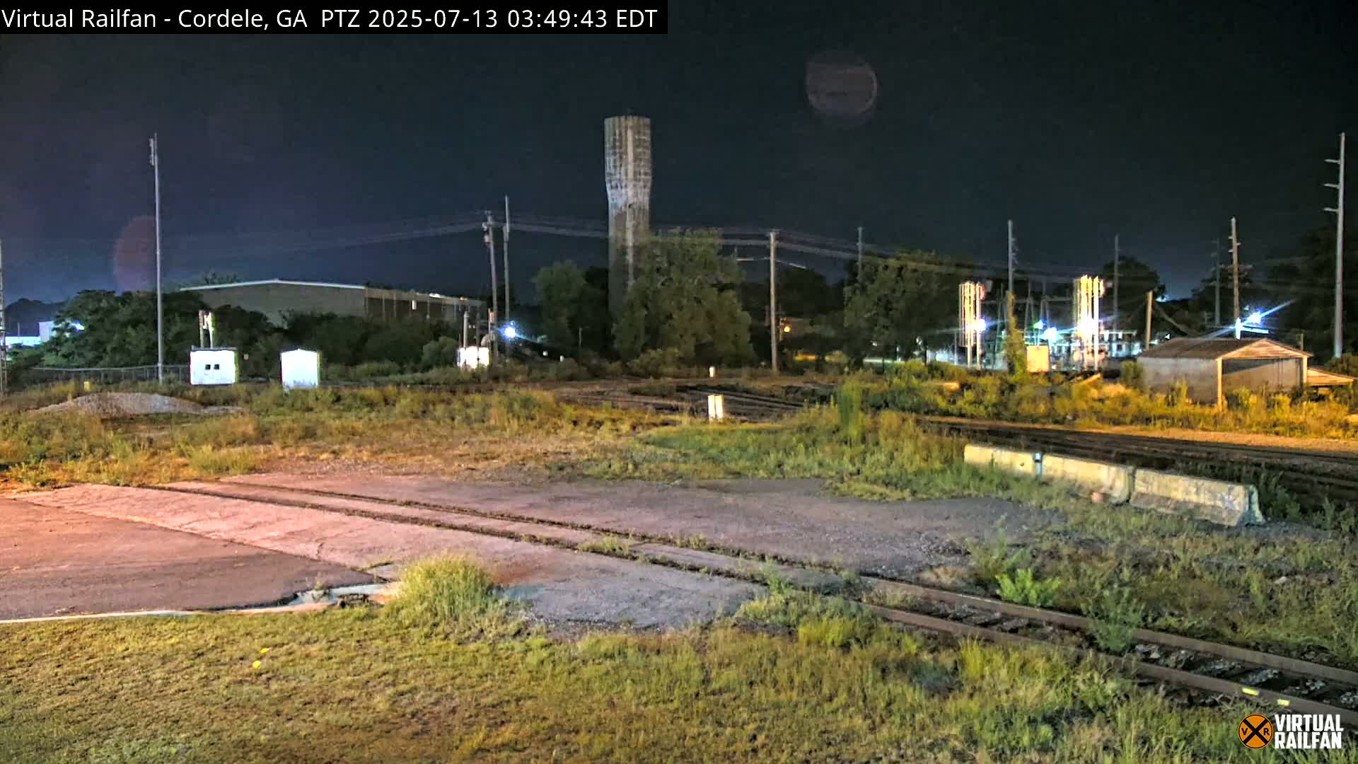 A nighttime view of a railway crossing with a tall tower and industrial buildings in the background under a clear, dark sky.