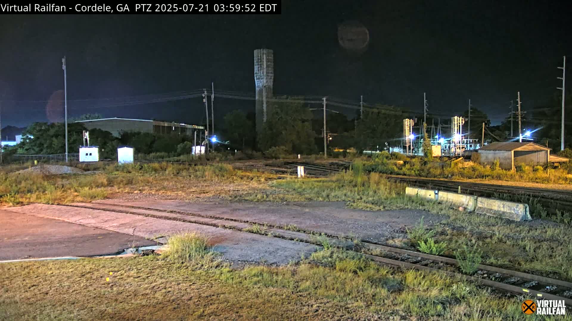 A nighttime view of railroad tracks and industrial buildings under a dark, clear sky, with a tall water tower visible in the background.