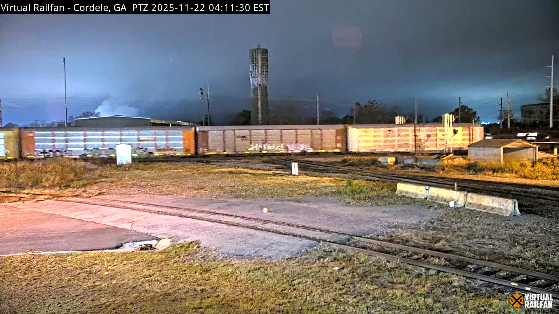 A brightly illuminated freight train moves through a dark, industrial rail yard at night under a cloudy sky, with multiple tracks, a tall concrete structure, and several distant buildings visible.