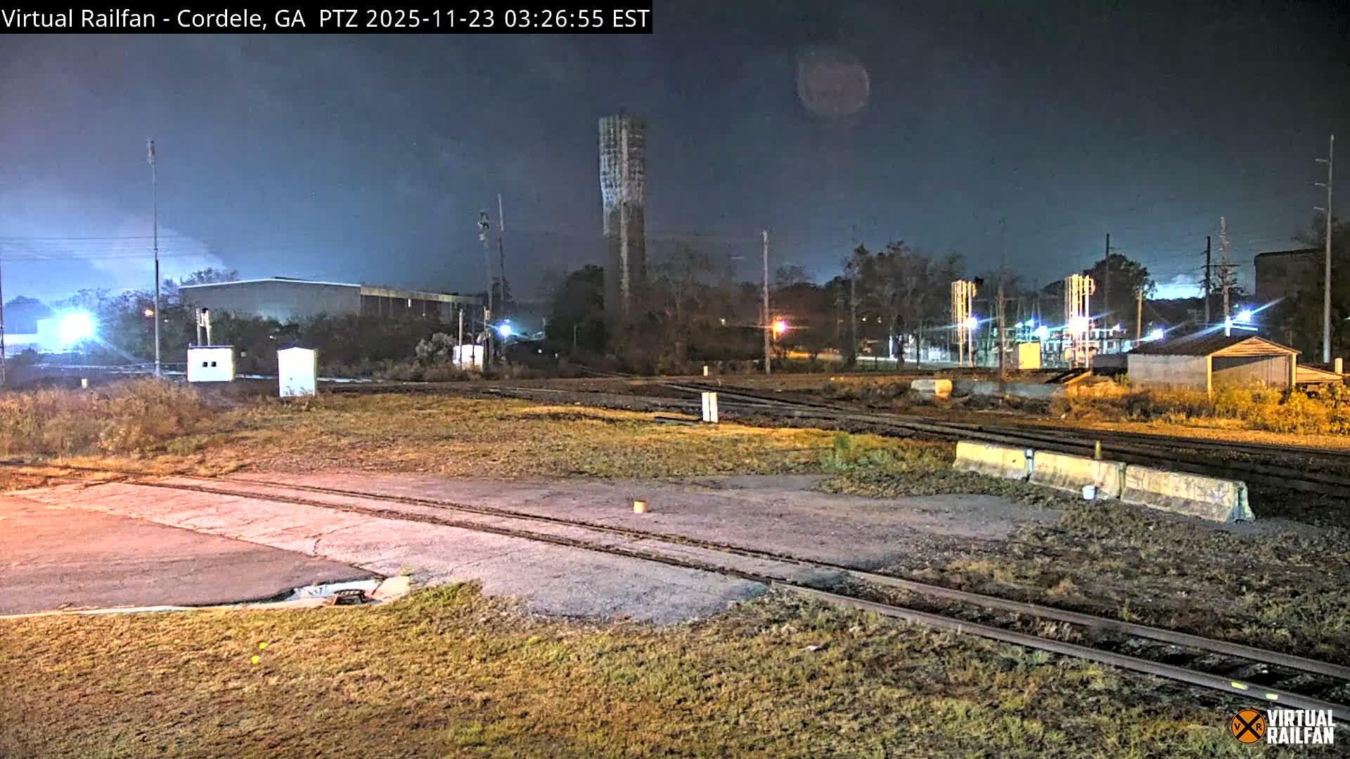 A dark, clear night reveals multiple railway tracks crisscrossing a grassy industrial area, with various buildings, power lines, and a tall concrete tower dimly illuminated by artificial lights in the background.
