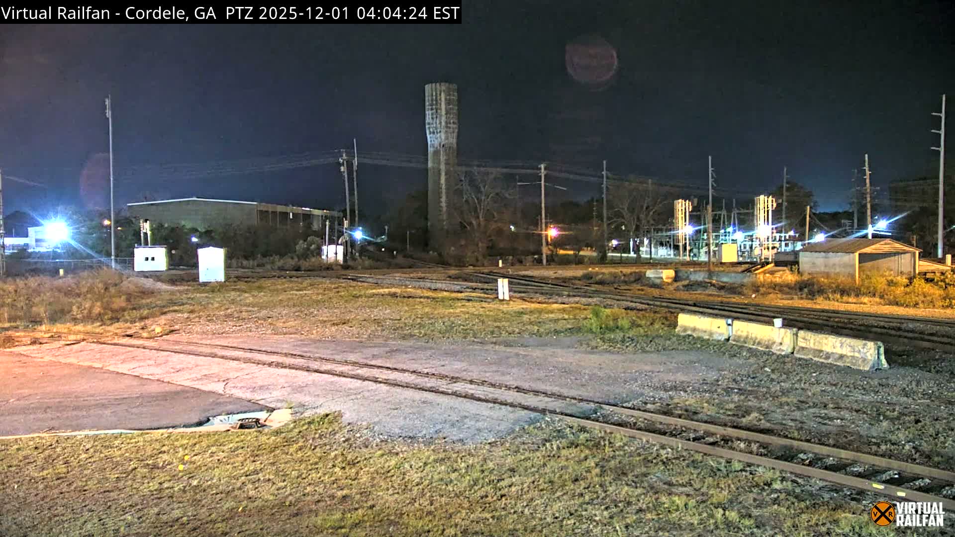 A clear night reveals a complex of railroad tracks crossing a paved area, flanked by a tall concrete tower, various industrial structures, utility poles, and a brightly lit electrical substation.
