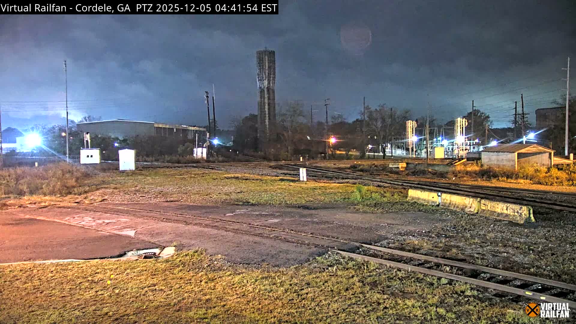 Multiple railroad tracks crisscross a grassy industrial area at night under an overcast sky, with a tall water tower, various buildings, and an illuminated electrical substation visible in the background.