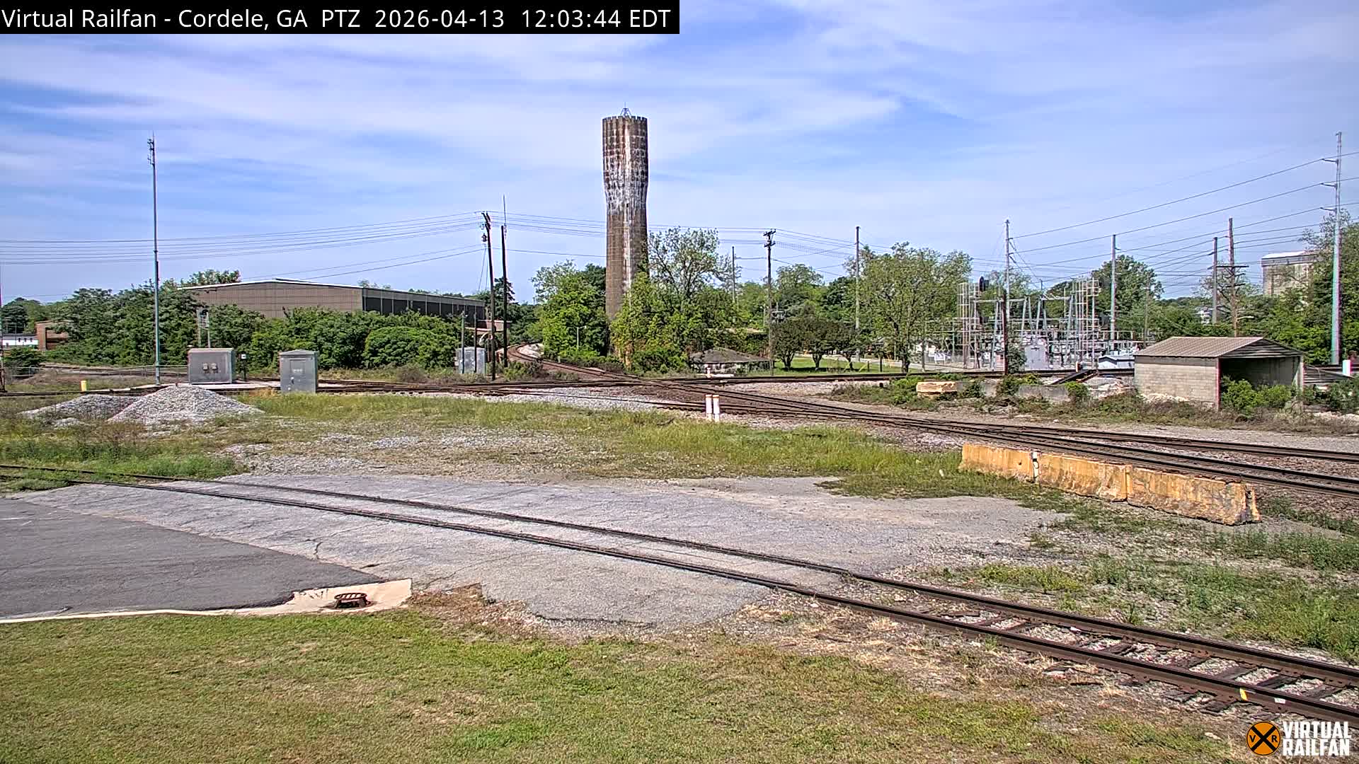 Multiple railroad tracks crisscross a grassy industrial area at night under an overcast sky, with a tall water tower, various buildings, and an illuminated electrical substation visible in the background.