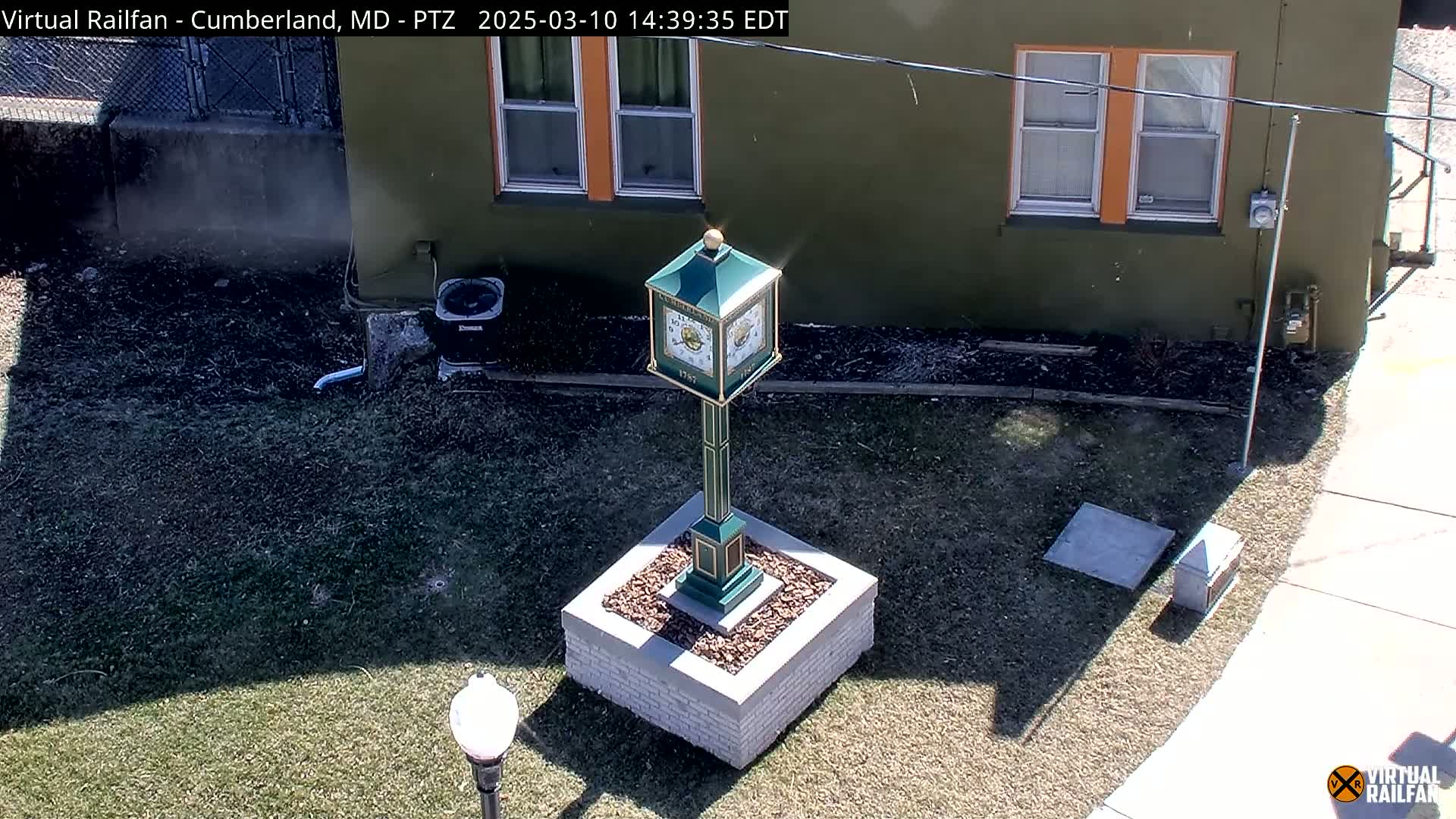 A green clock tower stands on a grassy area next to a building on a sunny day.