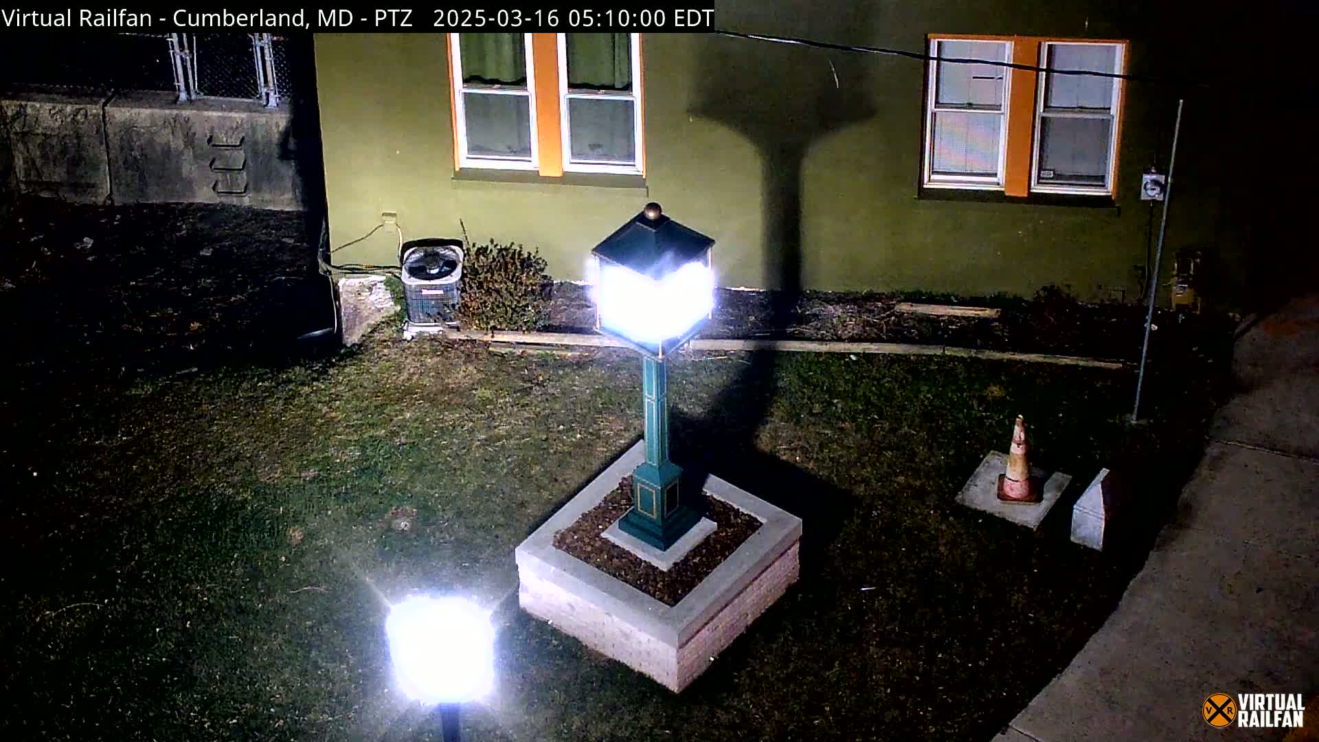 A brightly lit green lamp post stands on a small square base in a grassy area at night, near a building with orange-trimmed windows.