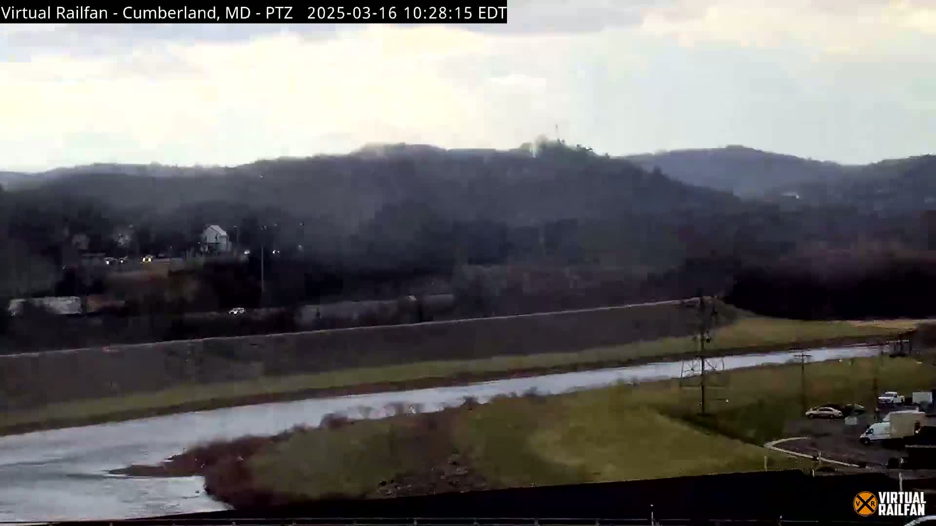 A wide shot shows a river flowing through a valley under an overcast sky, with hills and some vehicles visible in the background and a road and parking lot in the foreground.