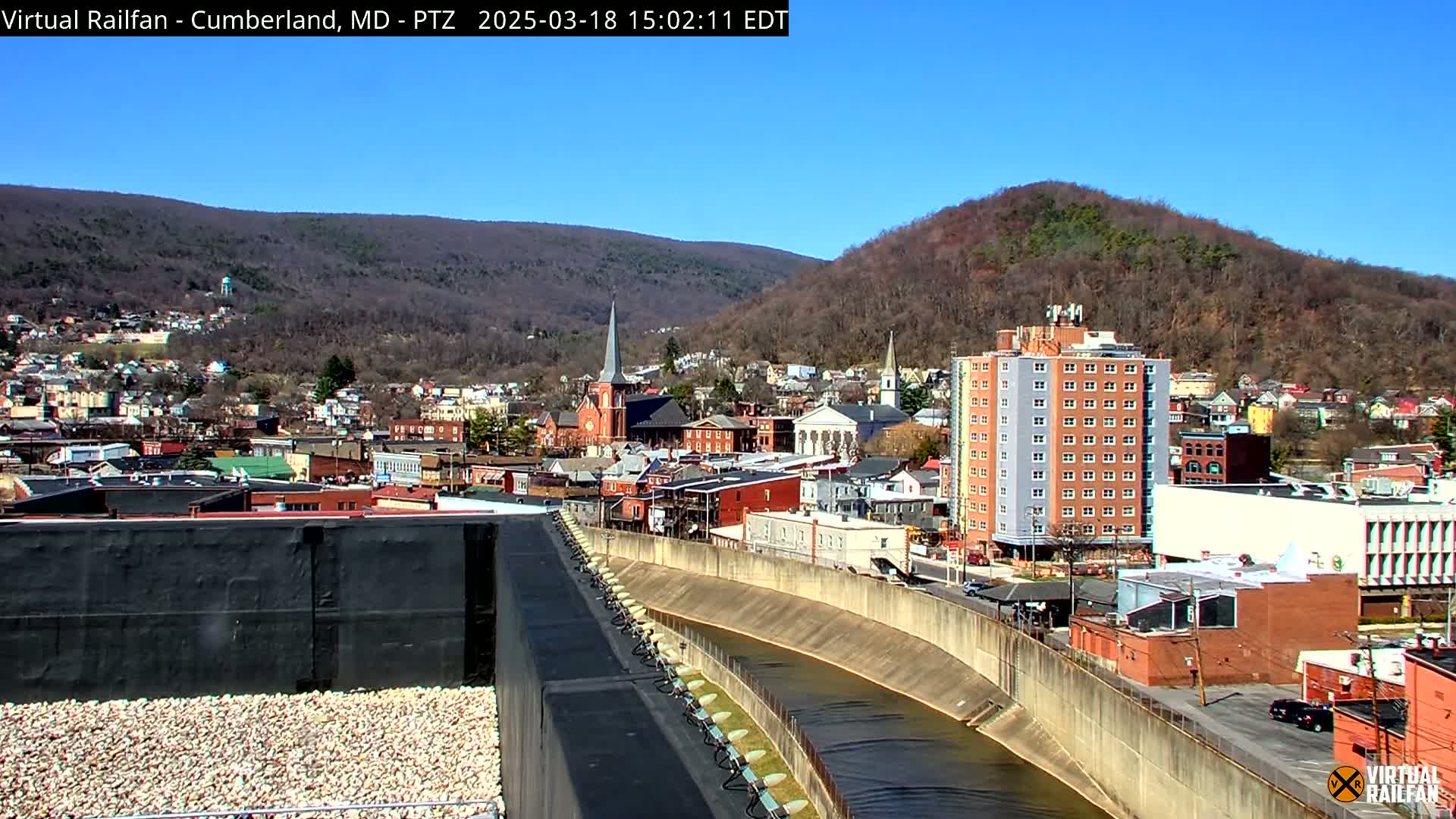 A clear, sunny day overlooks a town nestled in a valley between two hills, with a river curving through the town and a multi-story building visible near the riverbank.