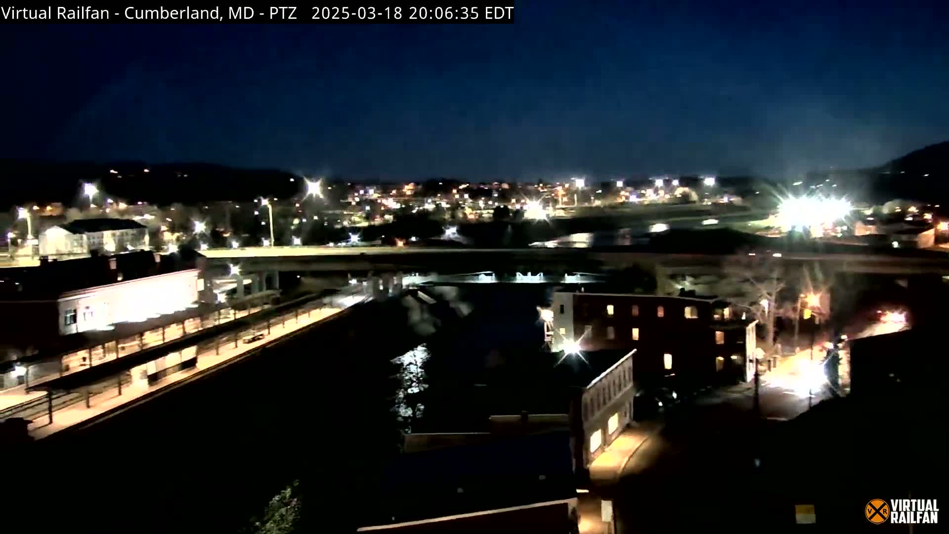A nighttime, elevated view of a city with a river, train station, and highway overpass under a clear, dark sky.