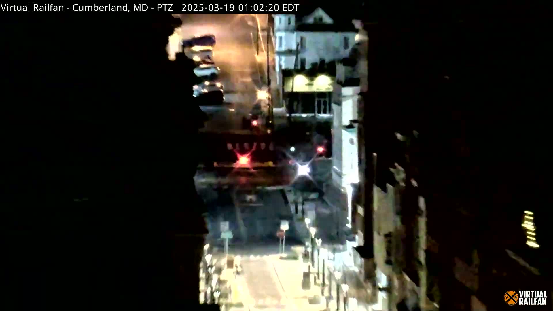A nighttime view of a street with buildings on either side, showing a train passing under a bridge.