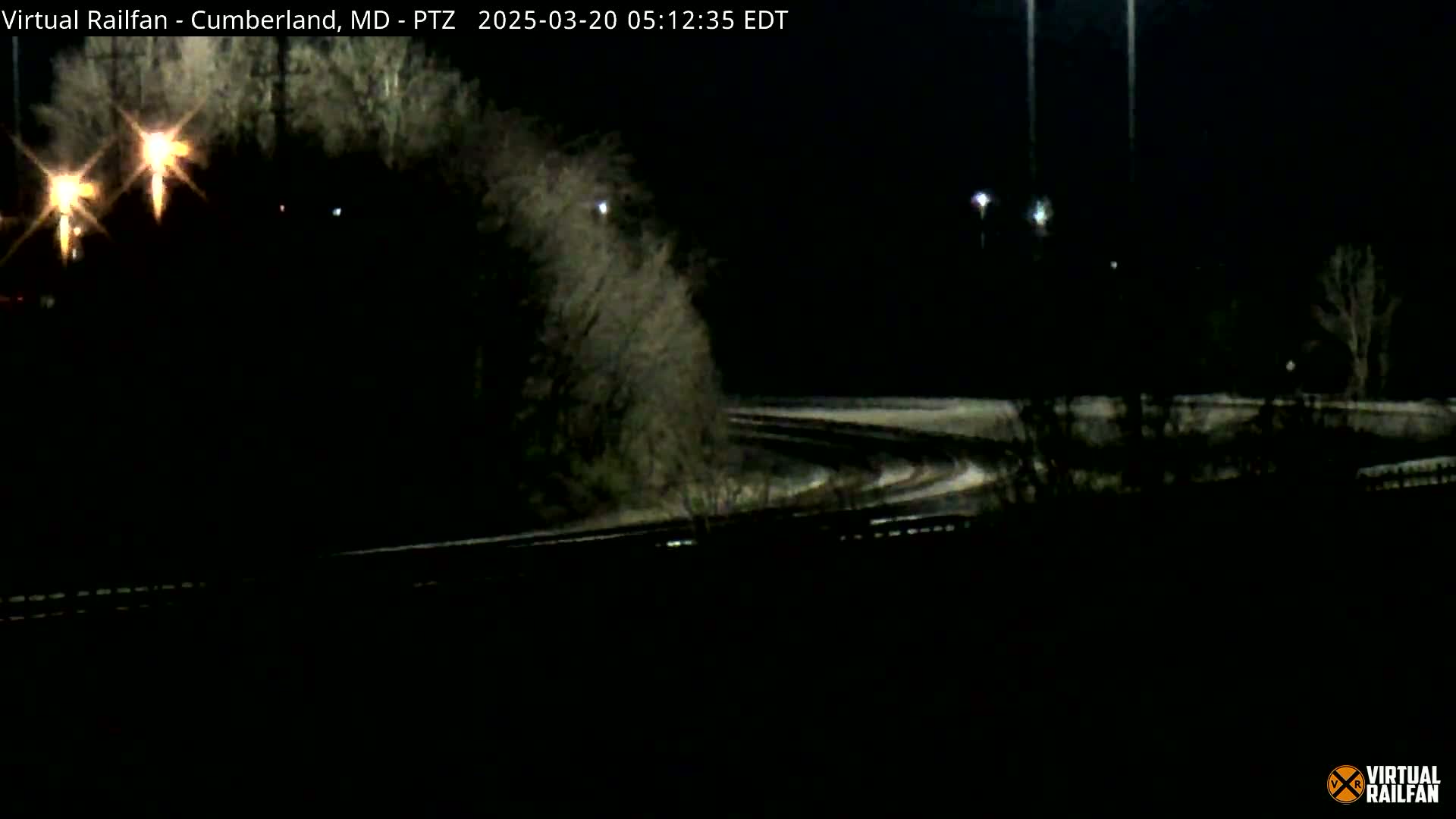 A dark, nighttime scene shows a curving road or railway track with sparse vegetation and distant lights under a clear sky.