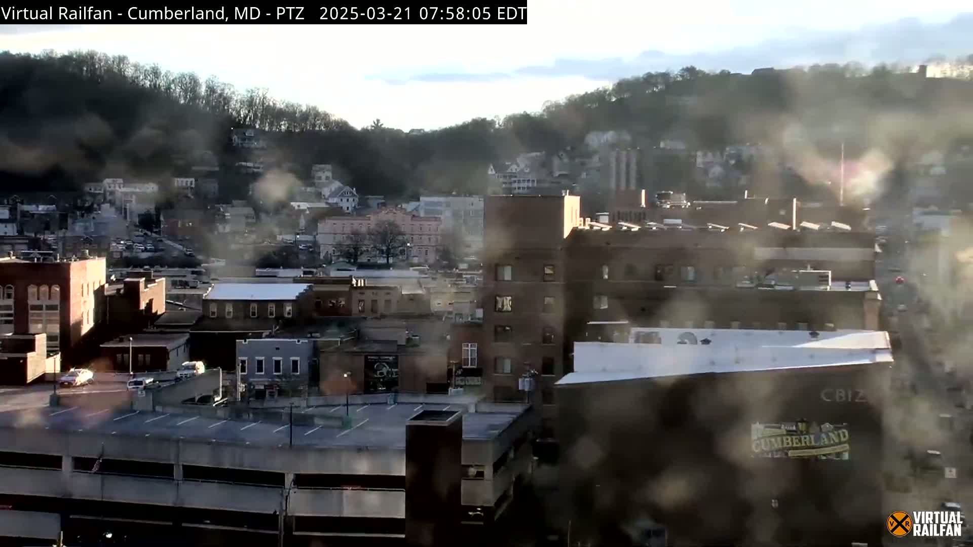 A hazy view of a city with brick buildings and a parking garage, situated on a hillside with trees in the background, under a partly cloudy sky.