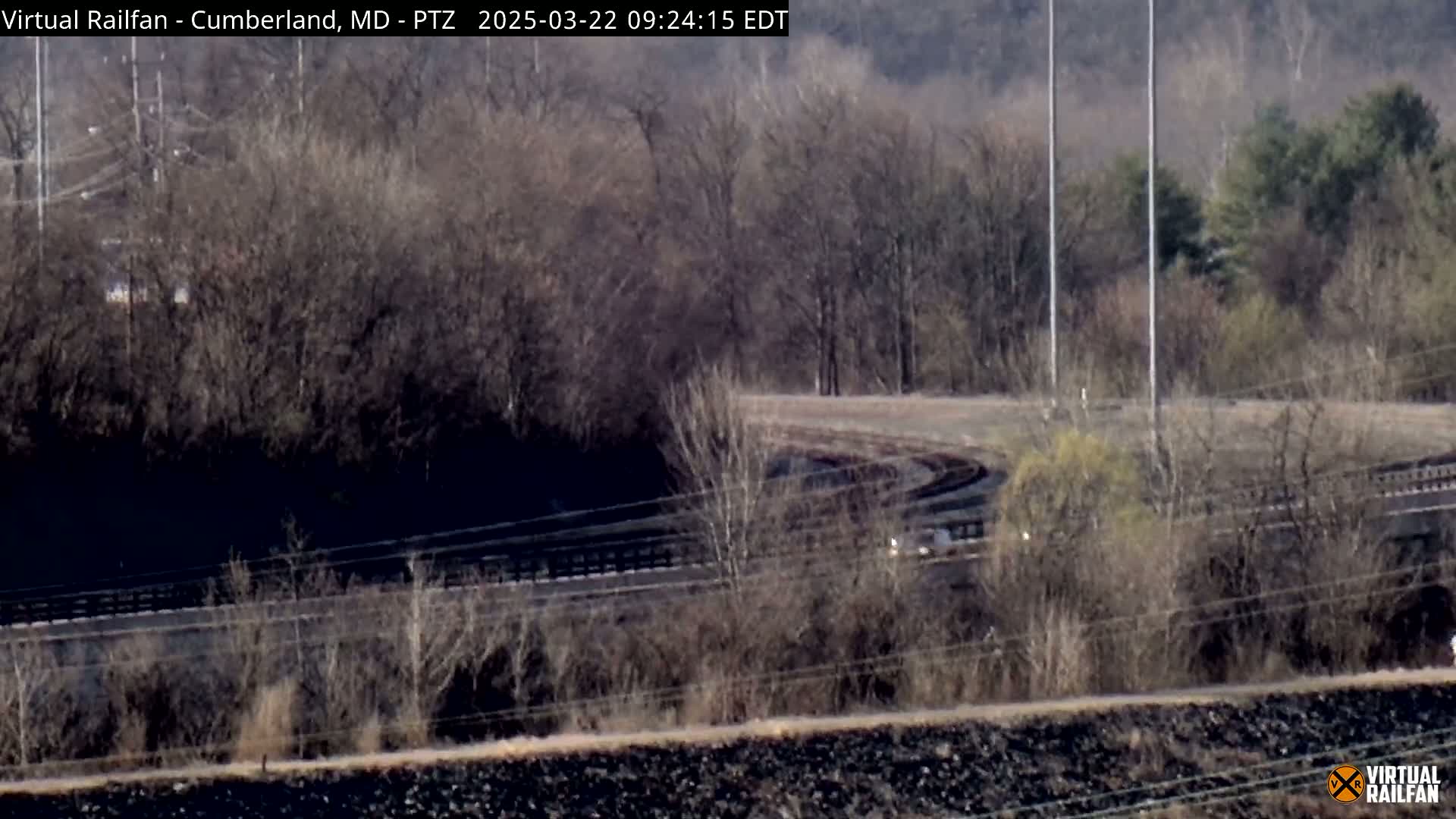 A view of a curving railway track partially obscured by bare trees and shrubs under a mostly sunny sky.