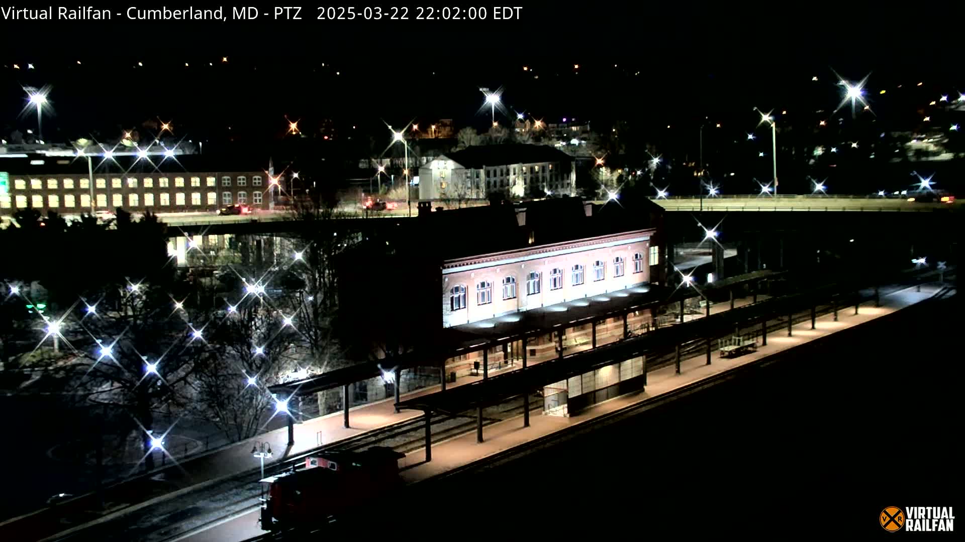 A nighttime, snow-dusted view of a train station with a long platform and a train partially visible, illuminated by streetlights and building lights.