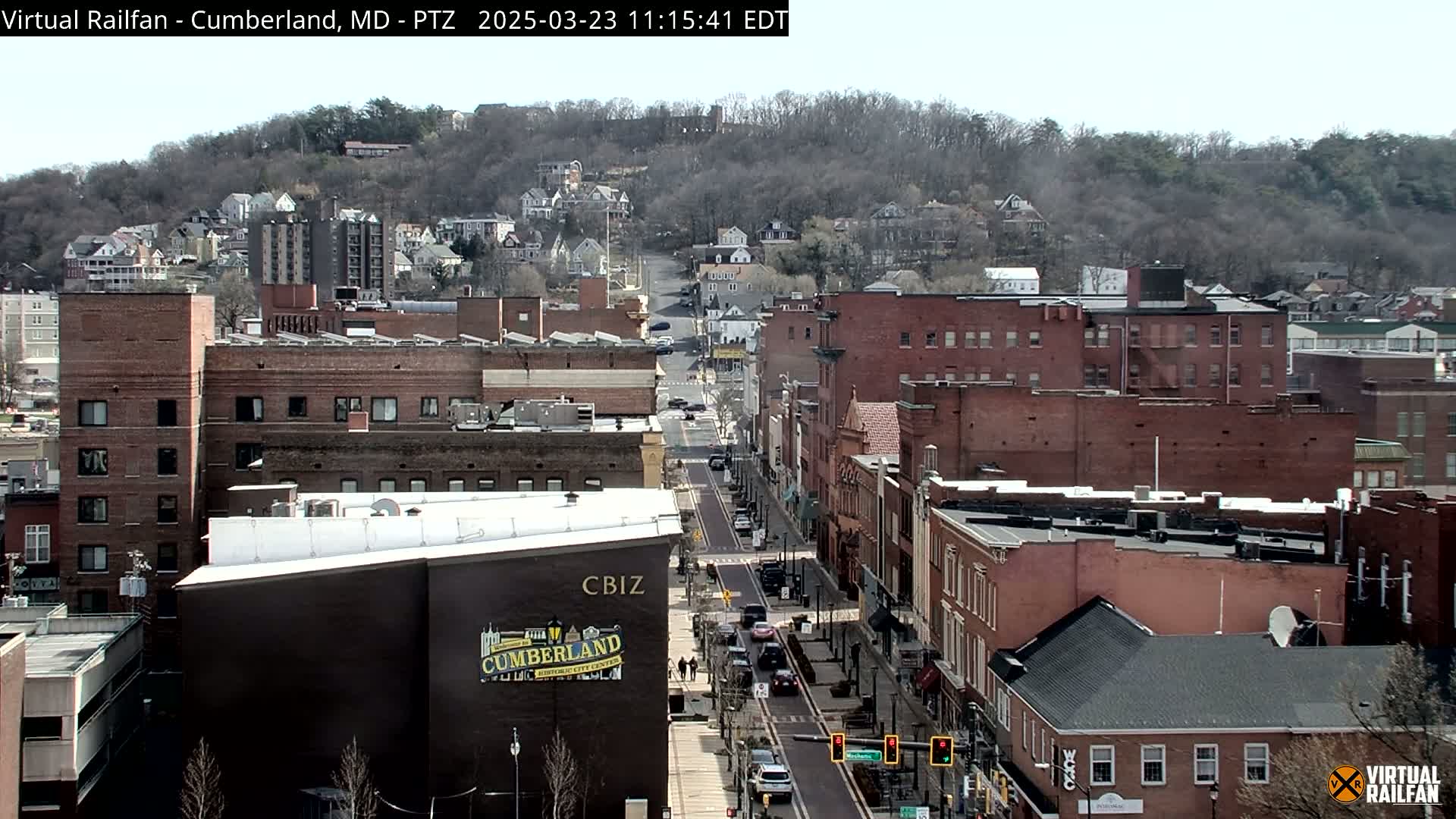 A mostly clear day reveals a long street in a city with brick buildings, hills in the background covered in leafless trees, and a few cars on the road.
