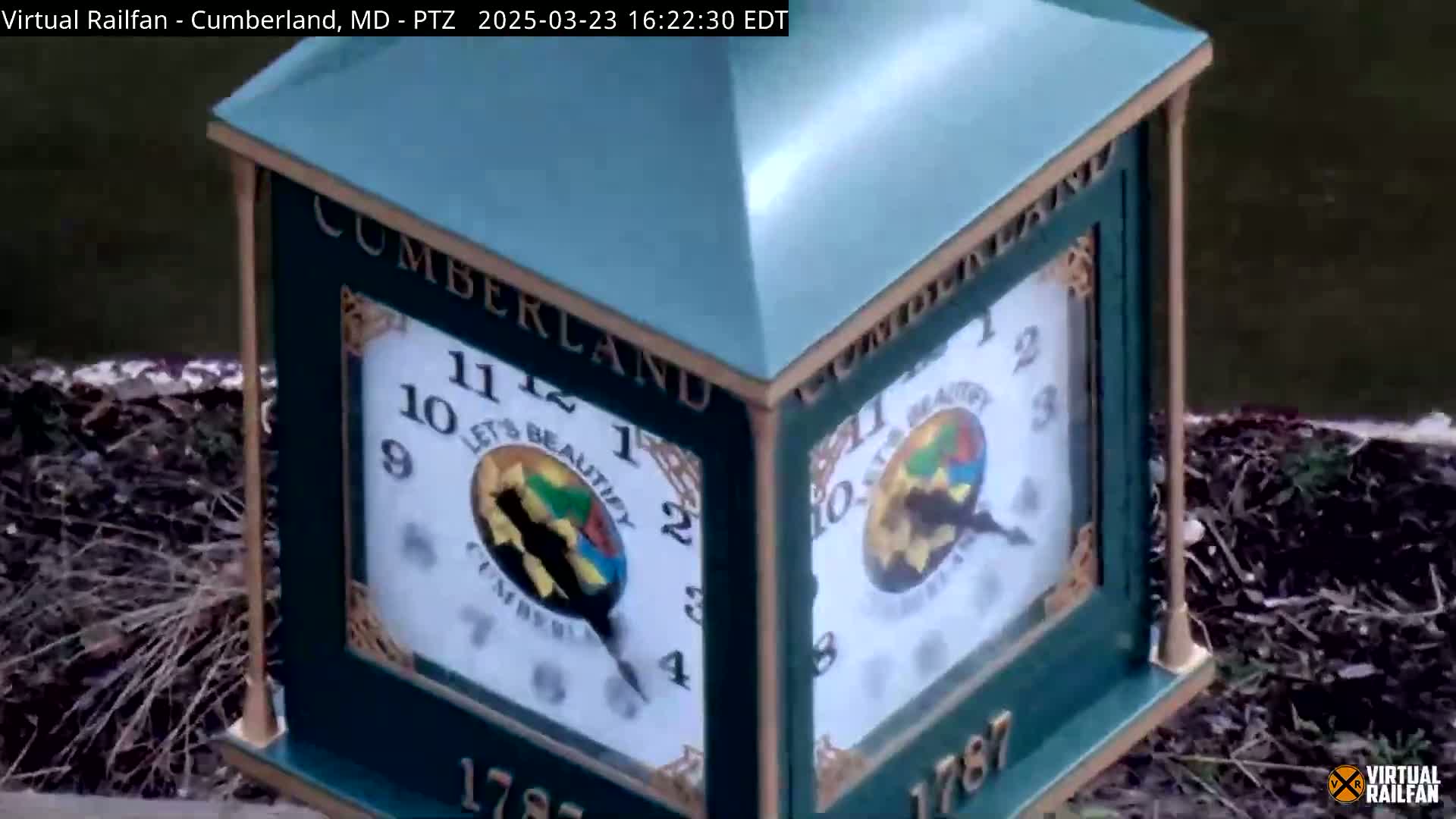 A green clock-like structure with two visible clock faces sits next to a body of dark water on a partly-covered ground.