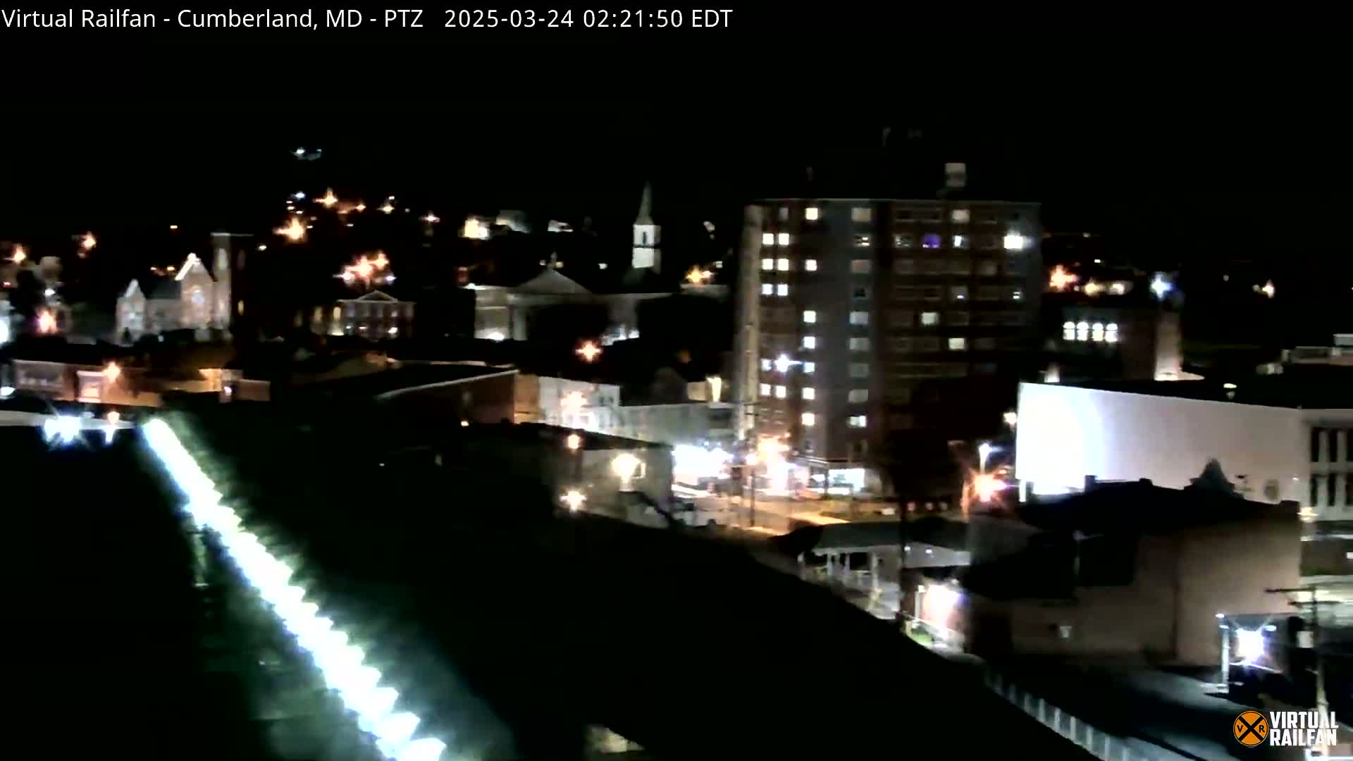 A nighttime long shot of a town, showing a line of lights along a roadway in the foreground and various buildings illuminated in the background.