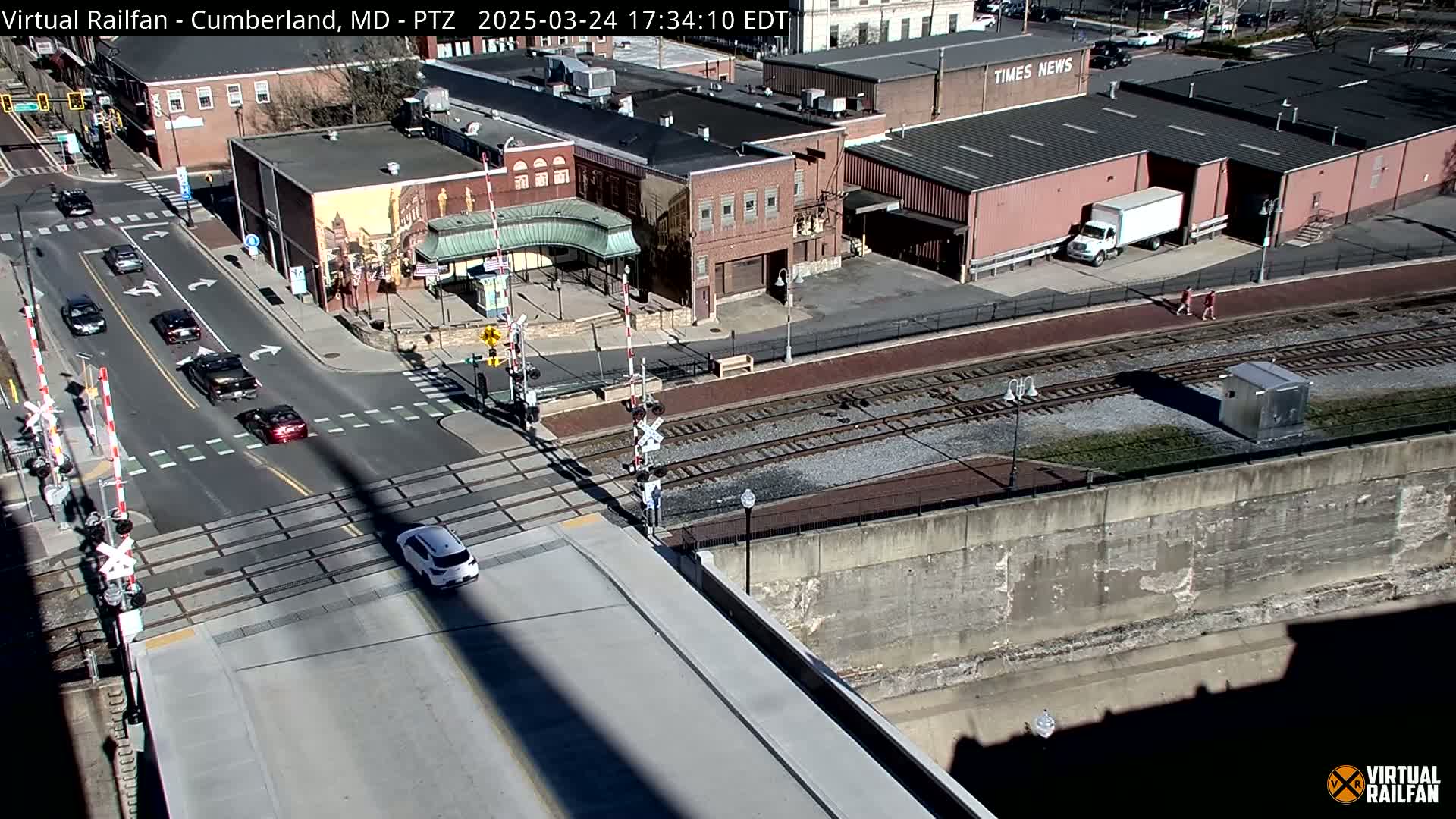 A sunny day view of a railroad crossing with several cars on the road and buildings in the background.