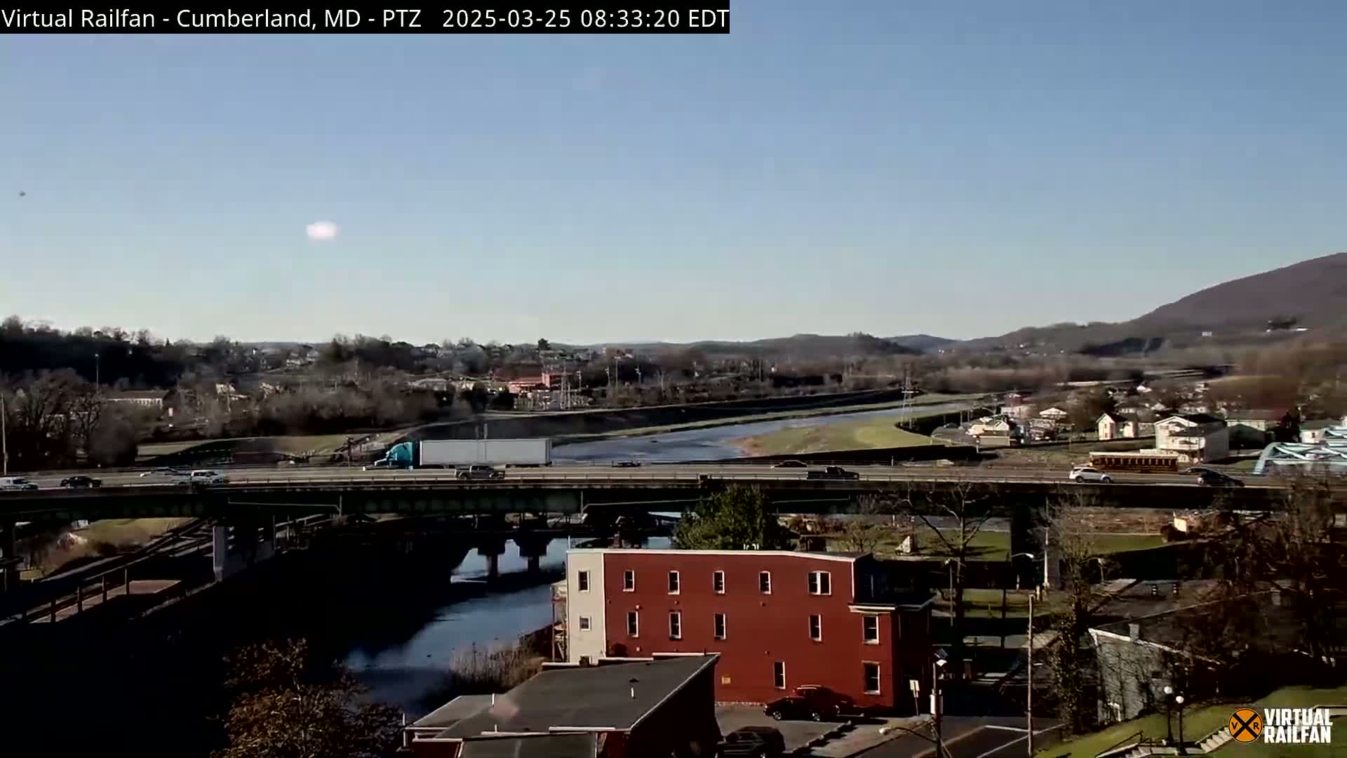 A highway overpass crosses a river, with a town and distant mountains visible under a clear, sunny sky.