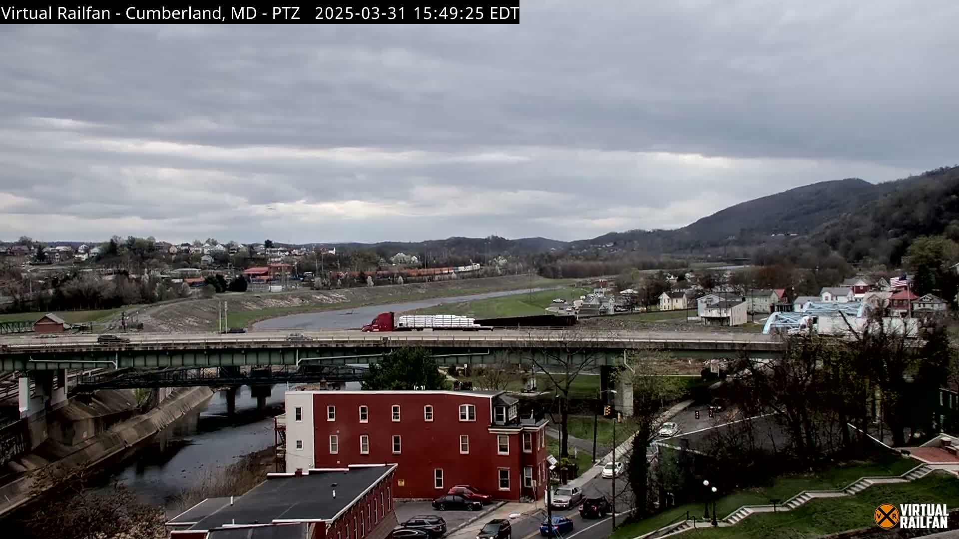 A semi-truck carrying a large load travels across a bridge over a river, with overcast skies and a town nestled between the river and a mountain in the background.