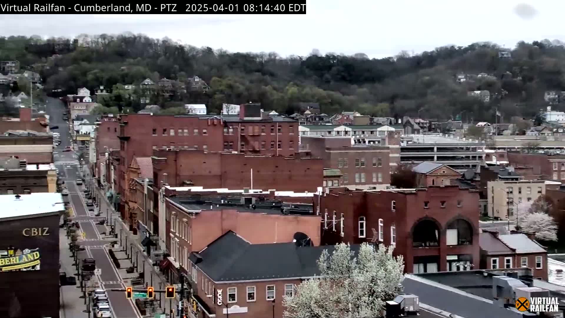 A mostly cloudy day reveals a long street lined with brick buildings, sloping up to a hill covered in trees.
