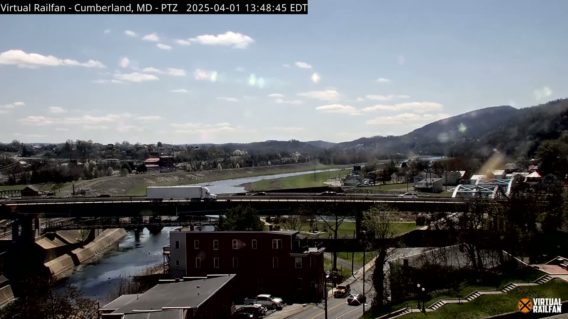 A partly cloudy day shows a river flowing under a bridge, with a town and hills in the background.