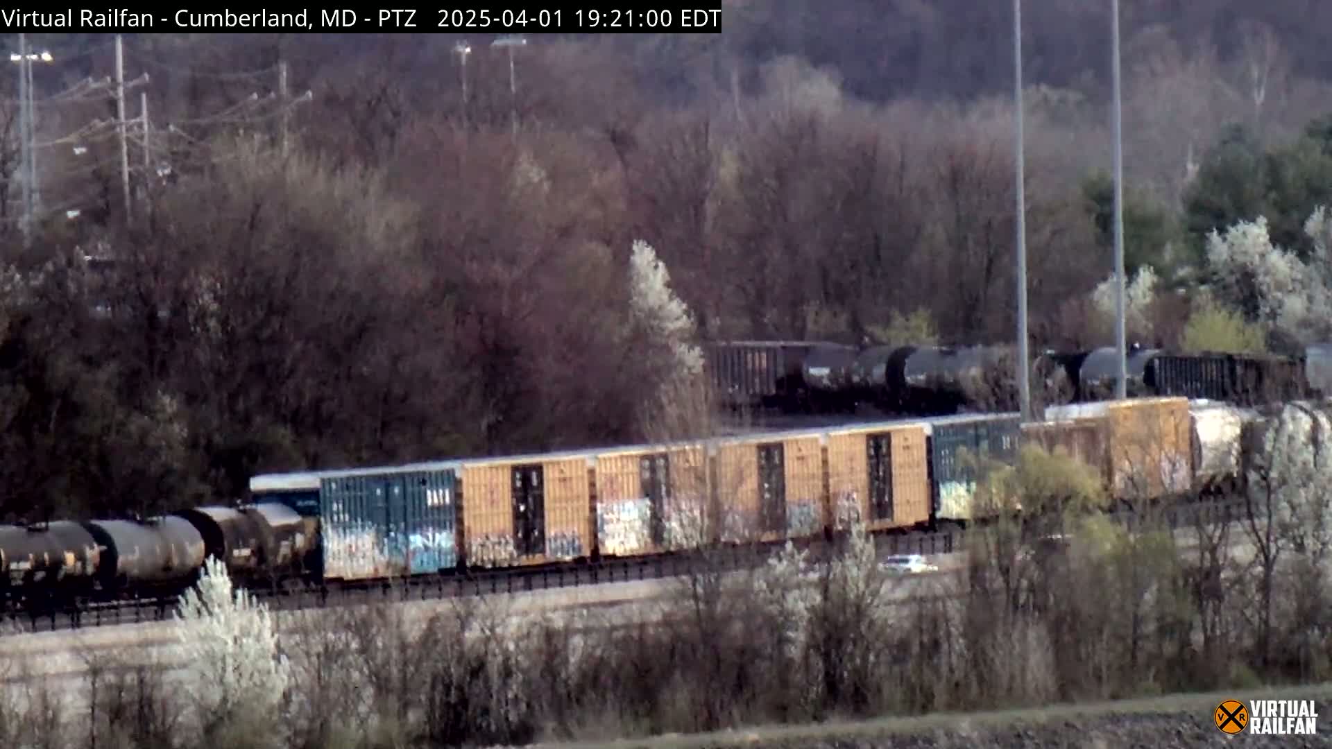 A long freight train, consisting of both tanker and box cars, travels through a wooded area on an overcast day.