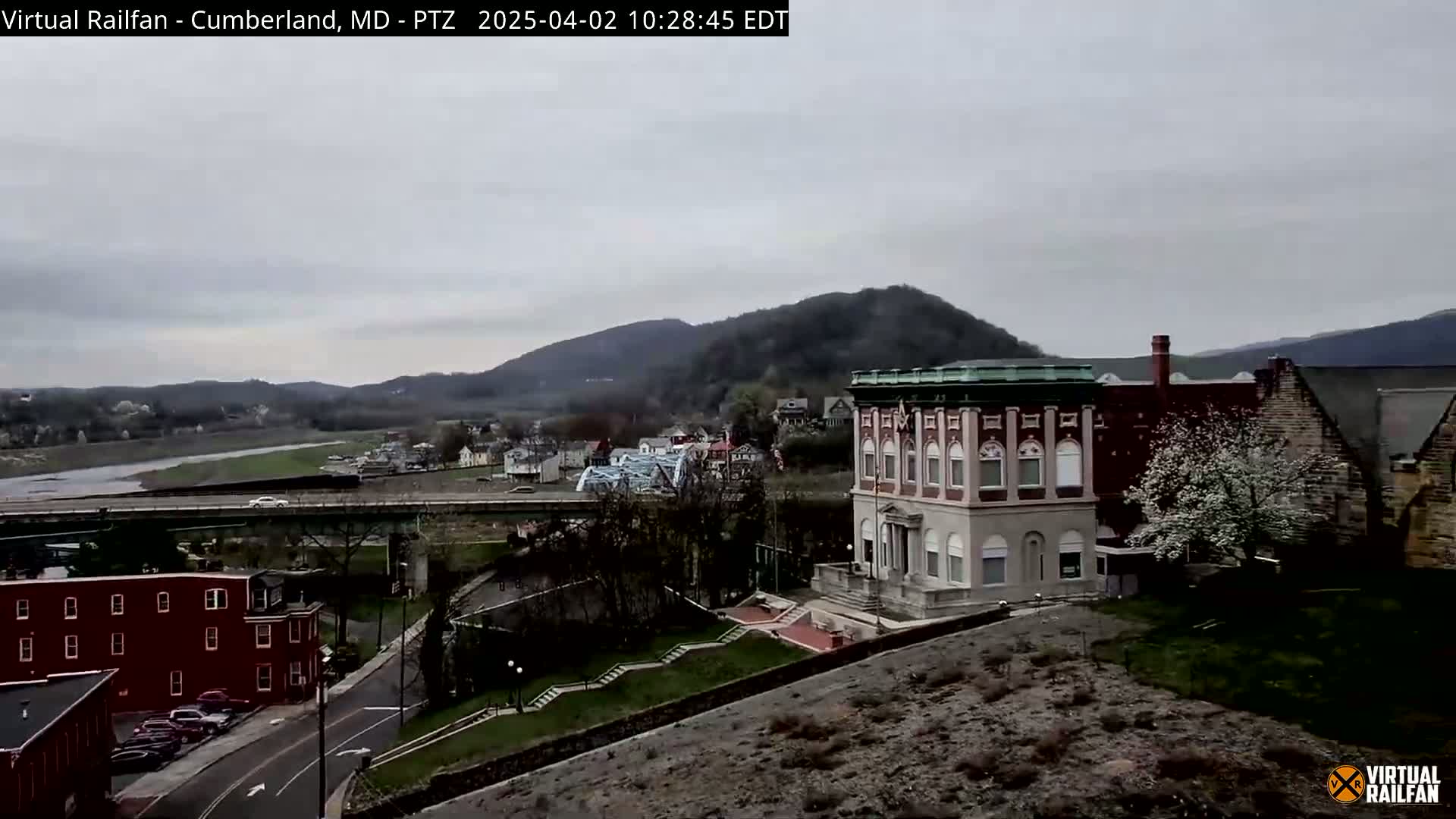 A view of a town situated beside a river, under an overcast sky, features a prominent brick building and hills in the background.