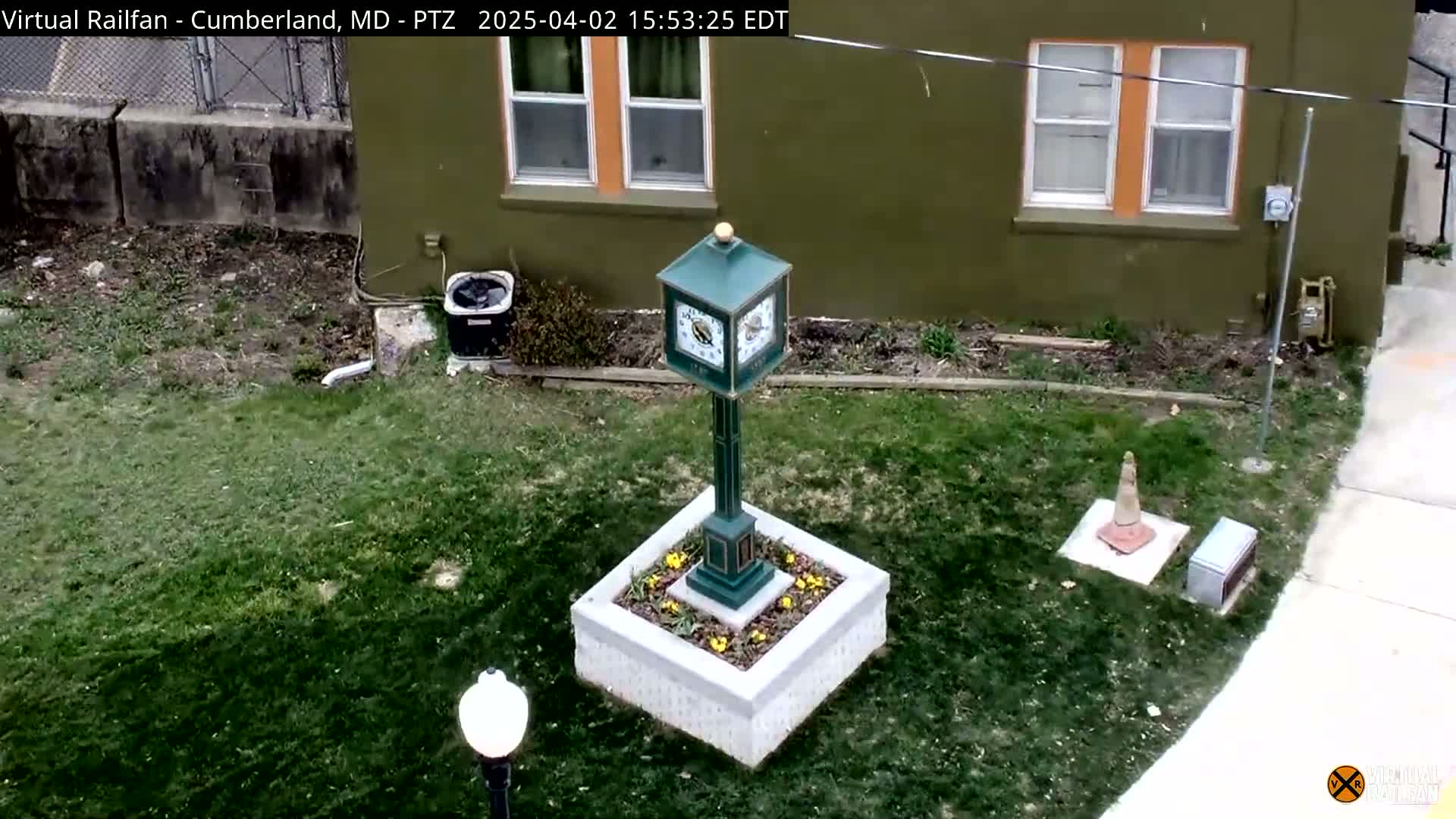 A green clock stands in a square planter surrounded by short grass next to a green building on a partly sunny day.