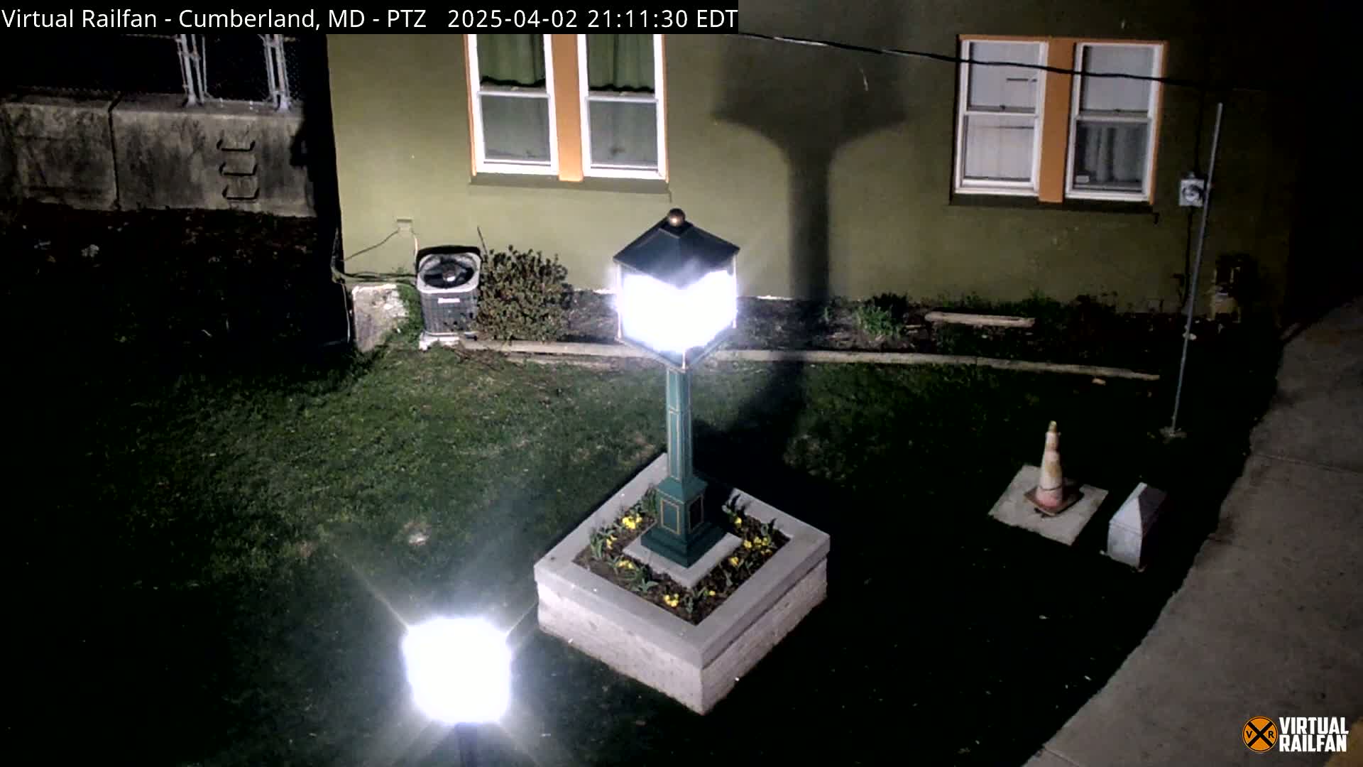 At night, a brightly lit lamppost stands in a small garden next to a building, with a traffic cone and other objects visible nearby.