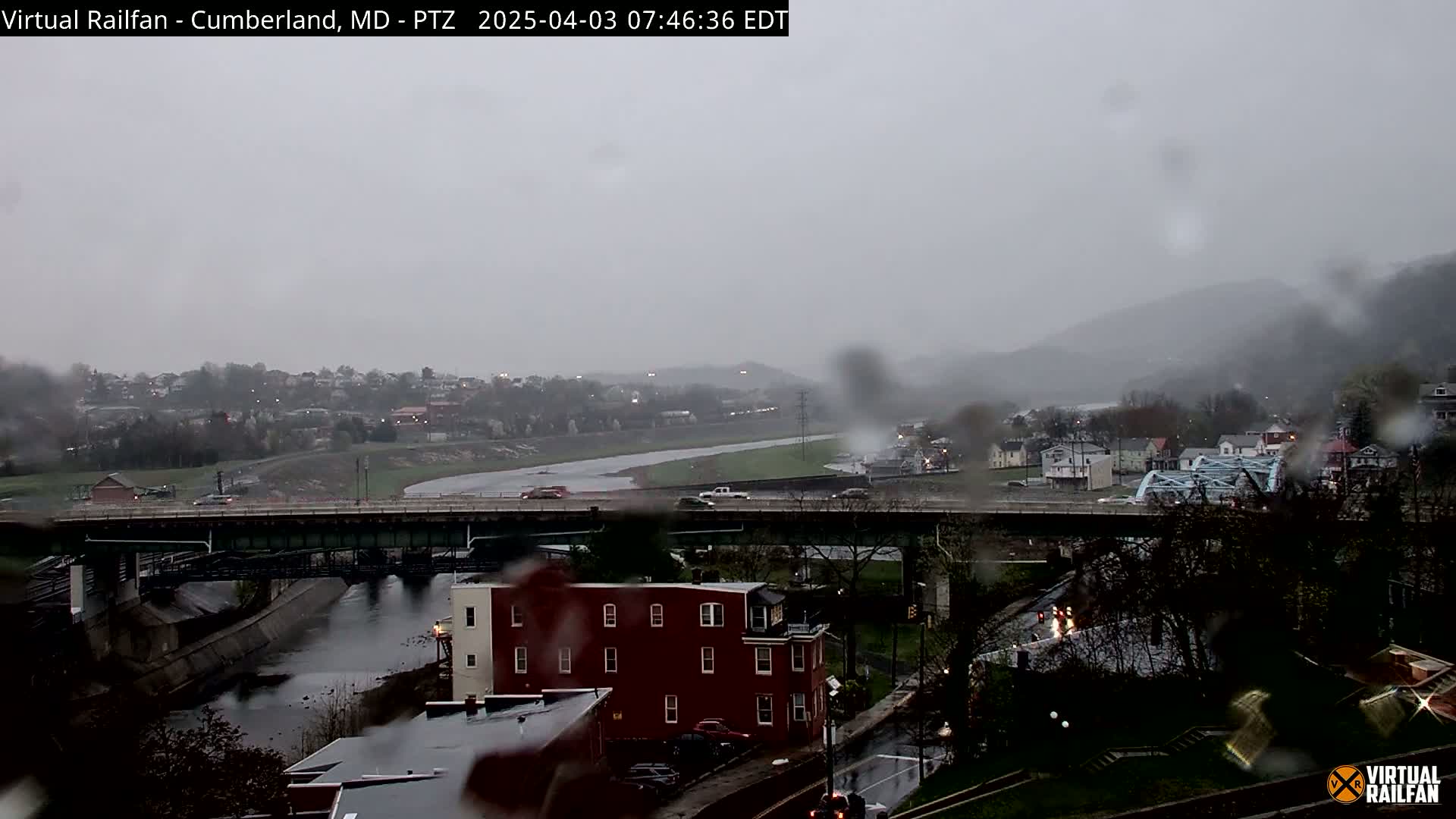 A rain-swept view of a river valley, featuring a bridge over a waterway, residential buildings, and a highway, all under a gray, overcast sky.