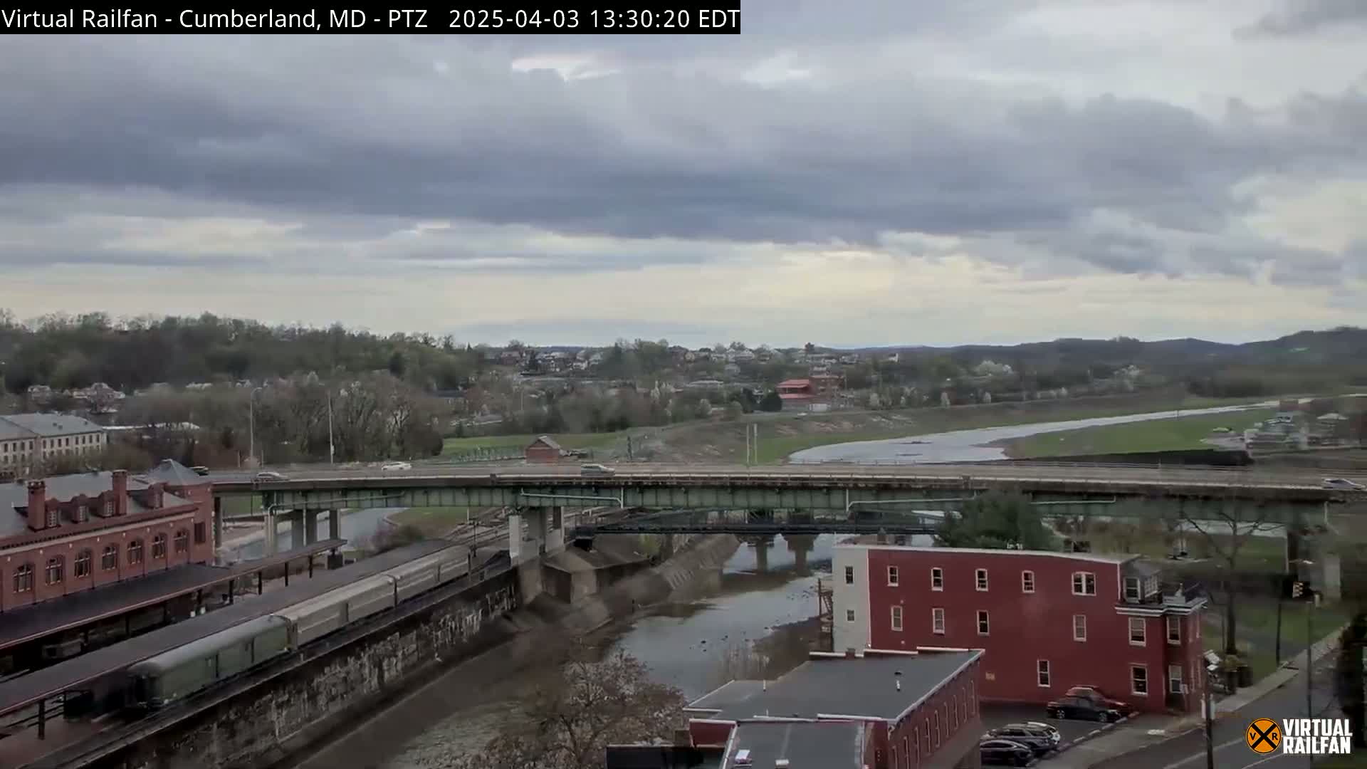 A long freight train sits in a station next to a river under an overcast sky, with a bridge and several buildings visible in the background.