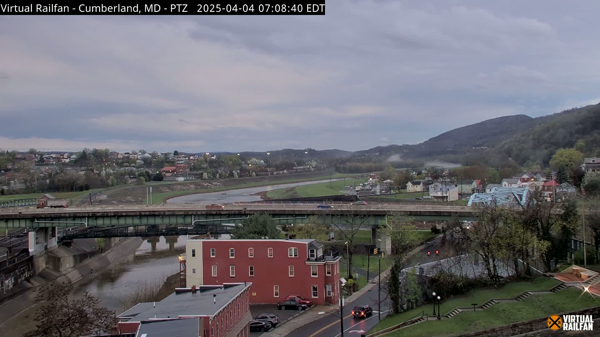 A river flows through a town nestled in a valley beneath a cloudy sky, with a bridge and buildings visible.
