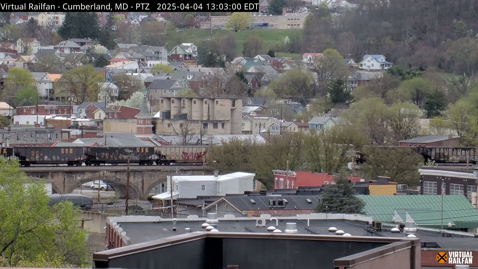 A freight train crosses a stone bridge over a street in a town with many houses and trees, under a mostly cloudy sky.