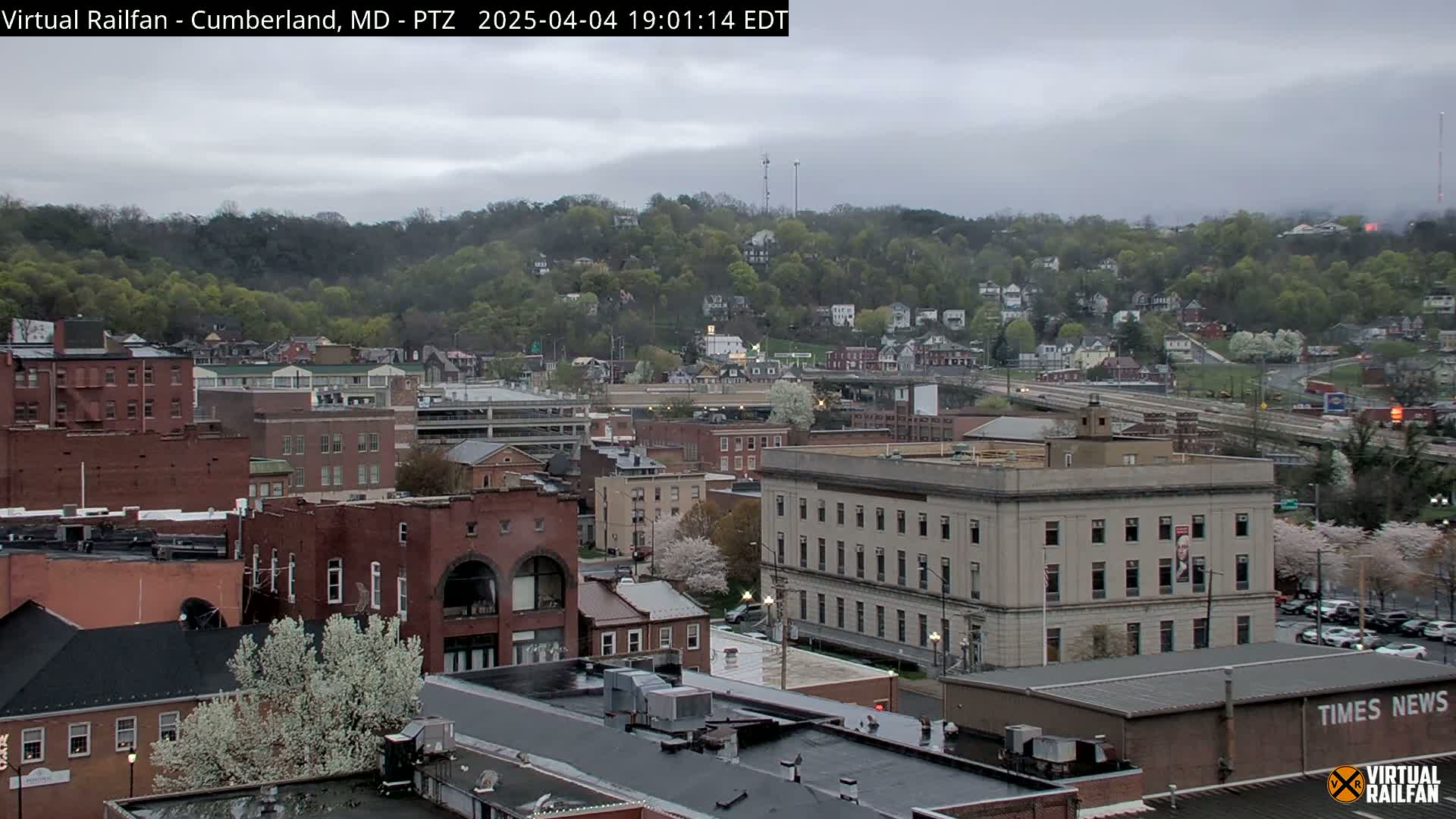A city with brick buildings and a hill covered in trees is seen under an overcast sky.