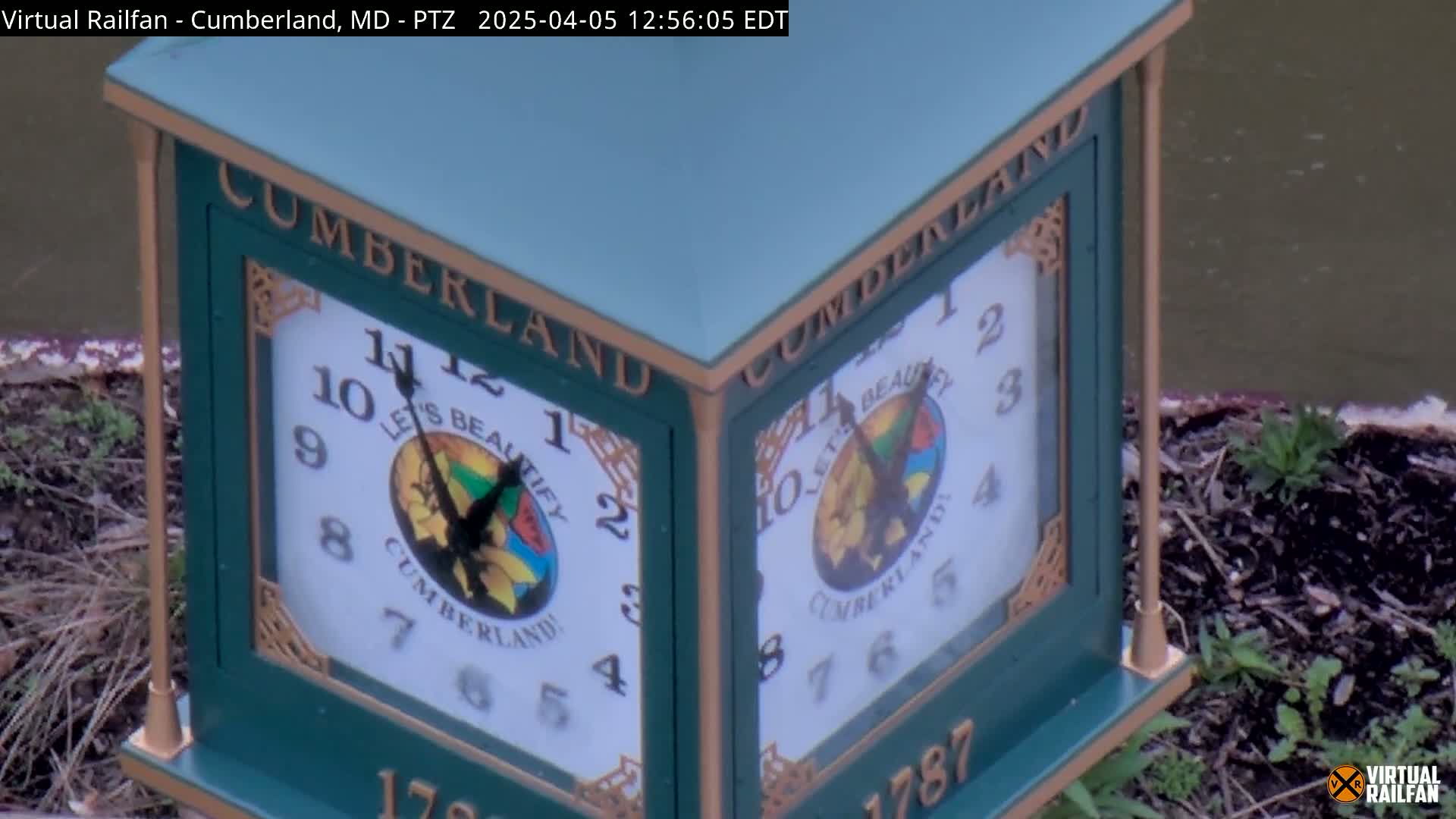 A green, multi-sided clock displaying the time, with the word "CUMBERLAND" on each face, sits beside a body of still water.