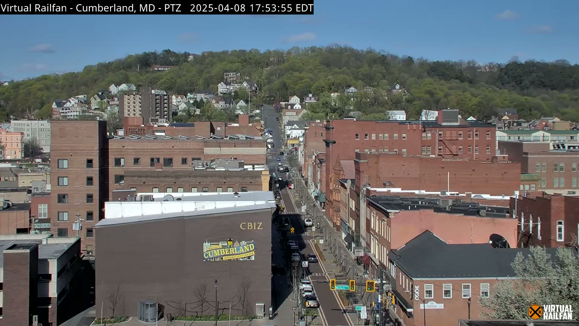 A mostly sunny view of a city street lined with brick buildings, some trees in bloom, and a hill covered in green foliage in the background.